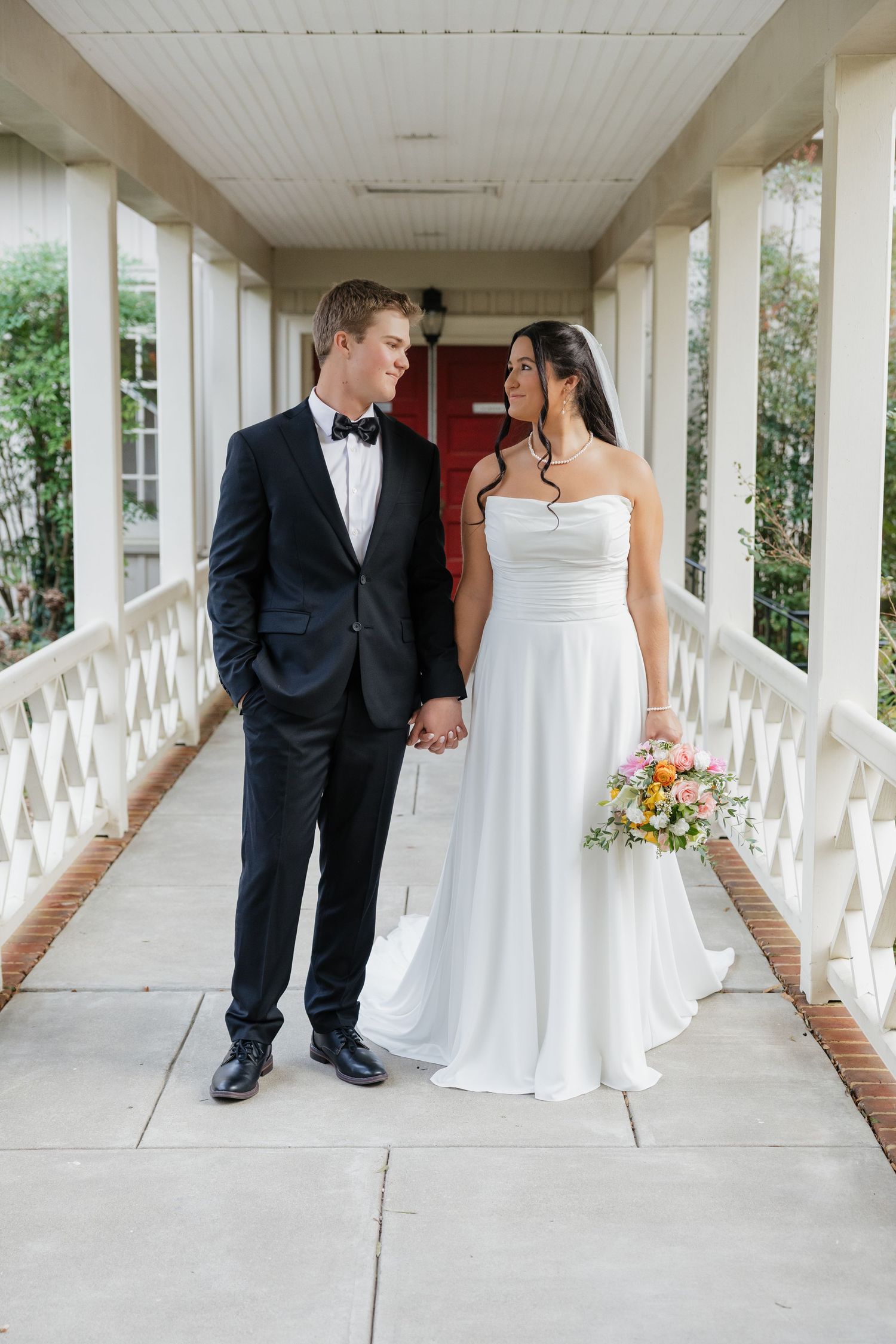 Bride and groom hold hands and gaze at each other while walking along a white columned wedding venue corridor.