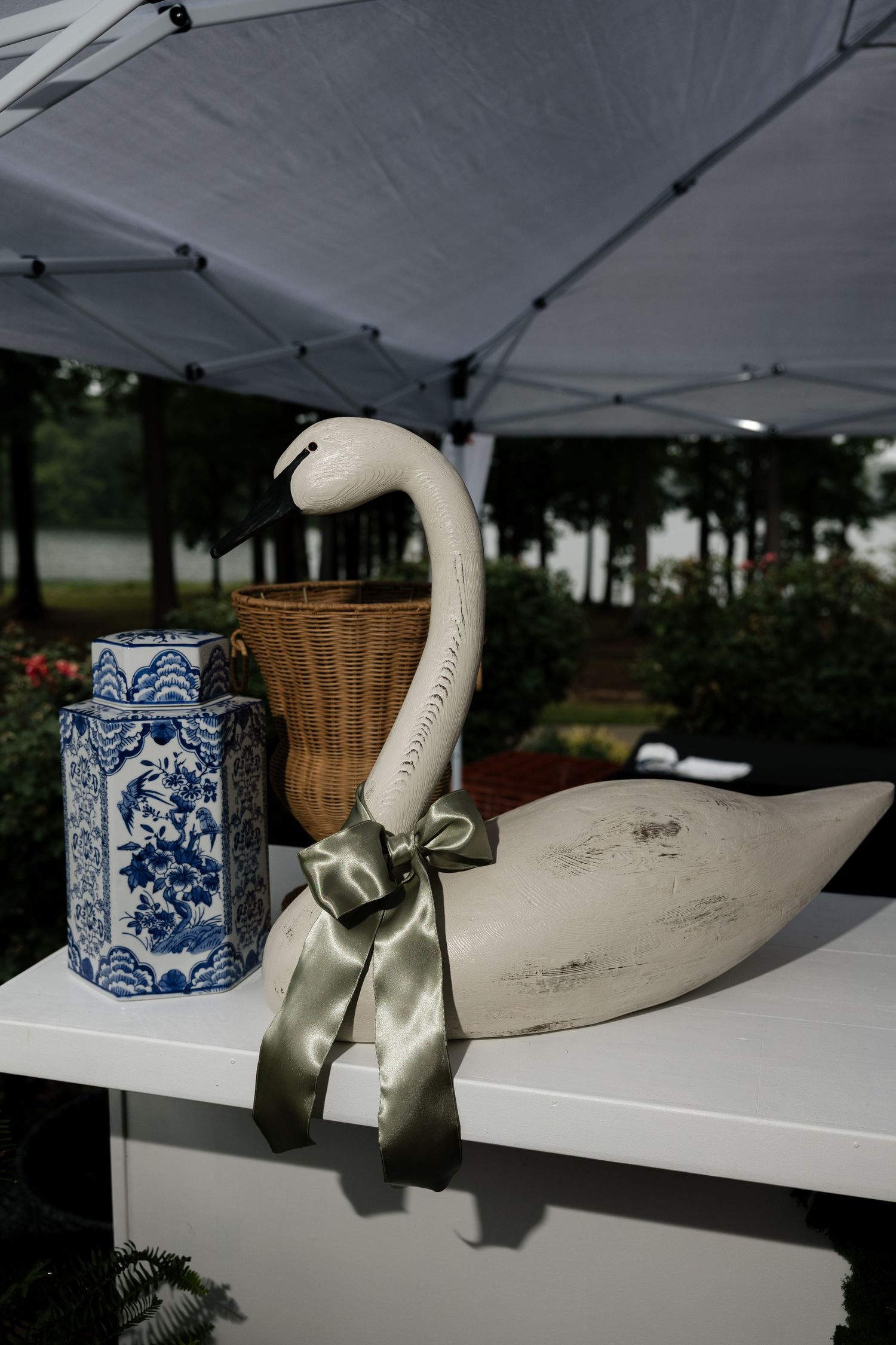 Decorative white wooden swan with sage green ribbon bow beside blue and white ceramic vase at outdoor event.