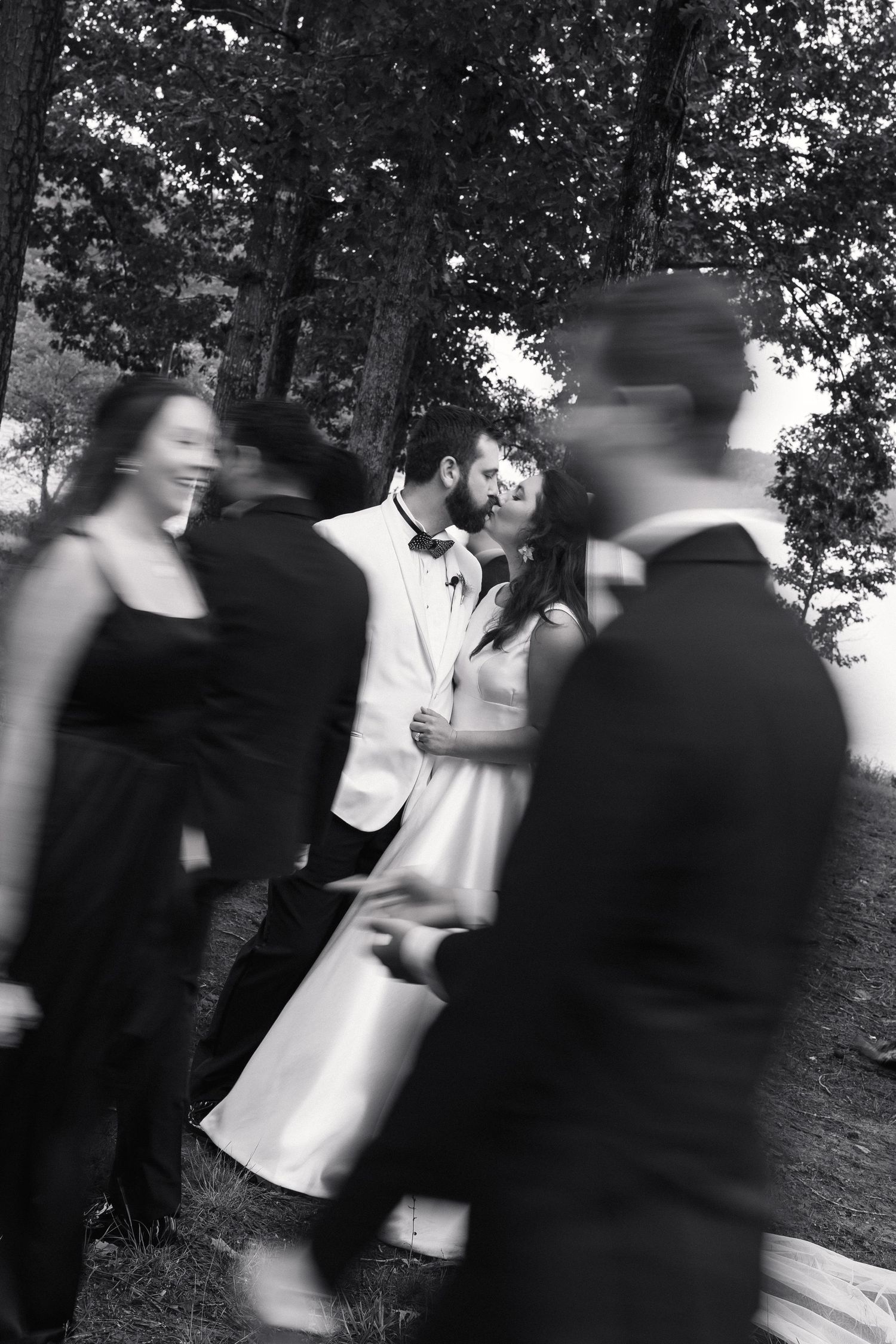 Black and white motion-blur photo of bride and groom kissing as wedding party moves around them.