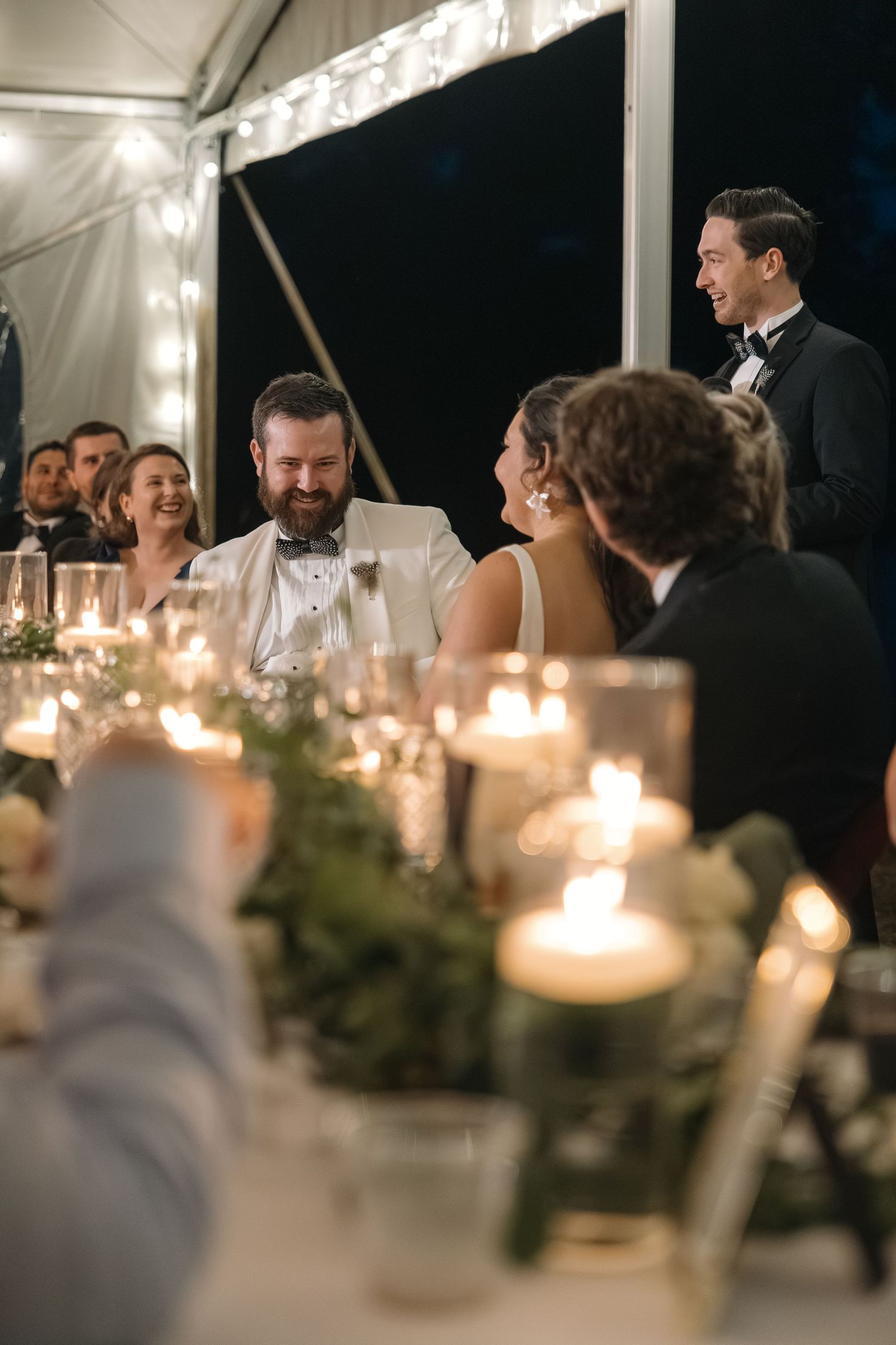 Groom in white tuxedo laughing with bride and guests at candlelit wedding reception head table.