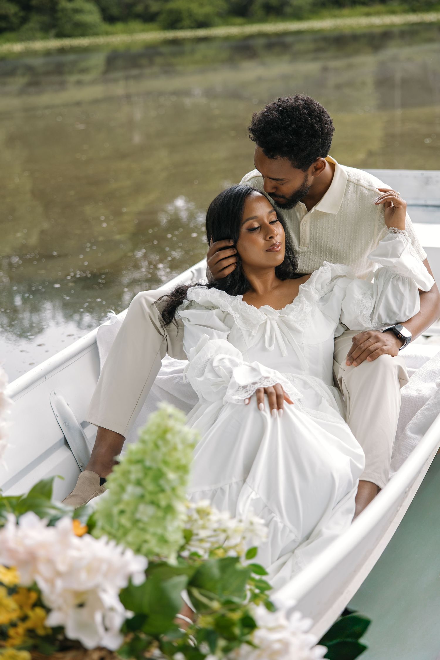 Romantic couple in white outfits sharing a tender moment on a boat near a serene lake, surrounded by flowers.