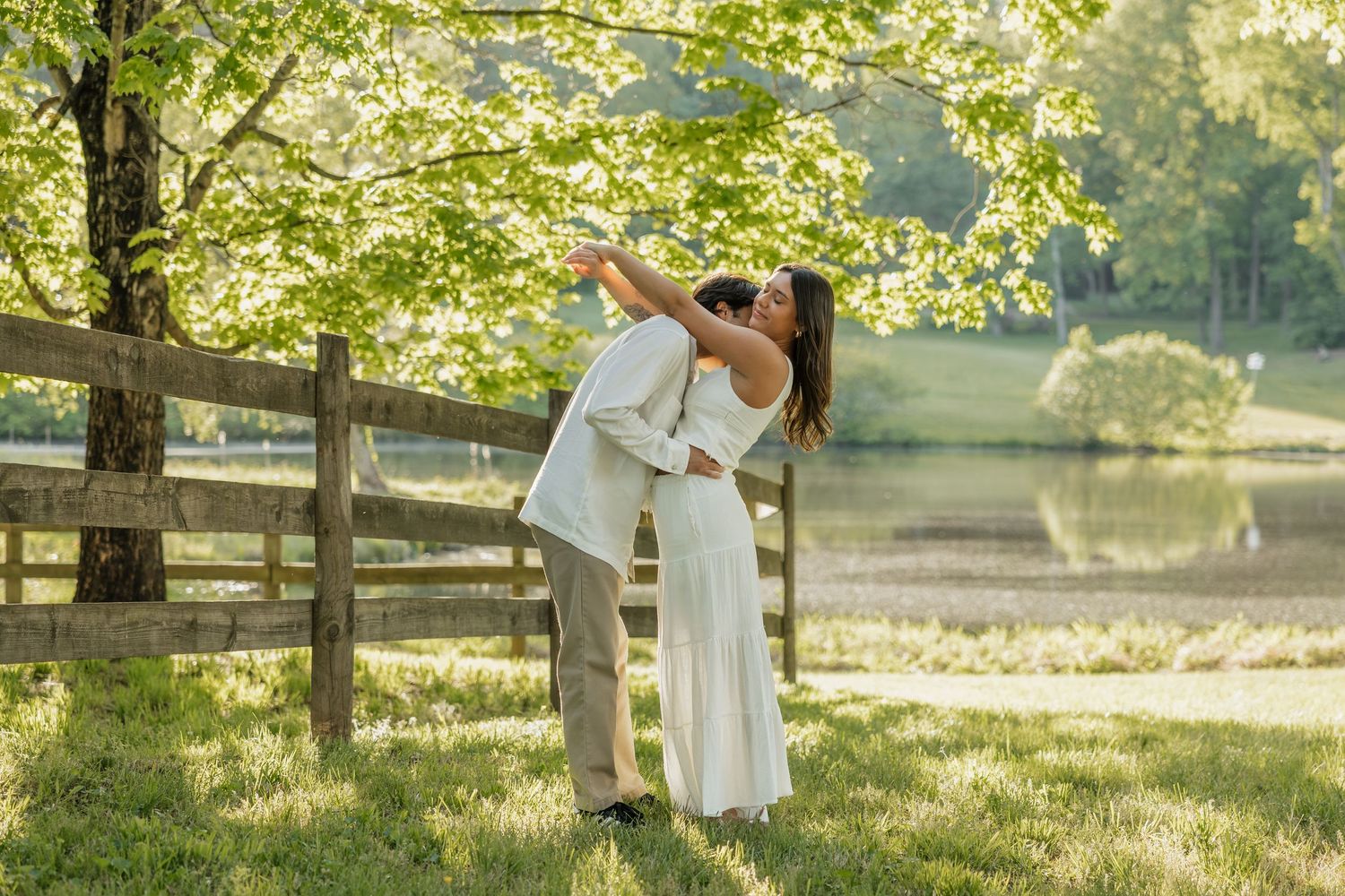 A couple shares a romantic dip kiss near a wooden fence with sunlit trees and a peaceful lake behind.