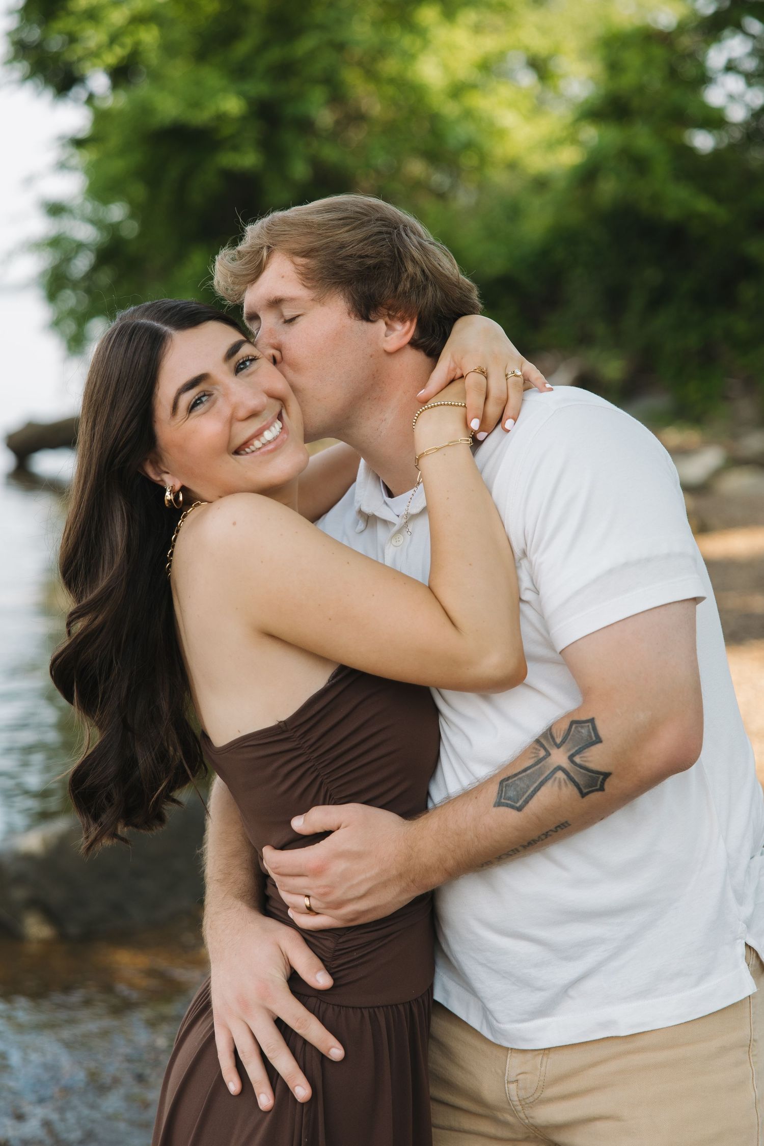 A happy woman smiles as her partner kisses her cheek while embracing near a scenic waterside.