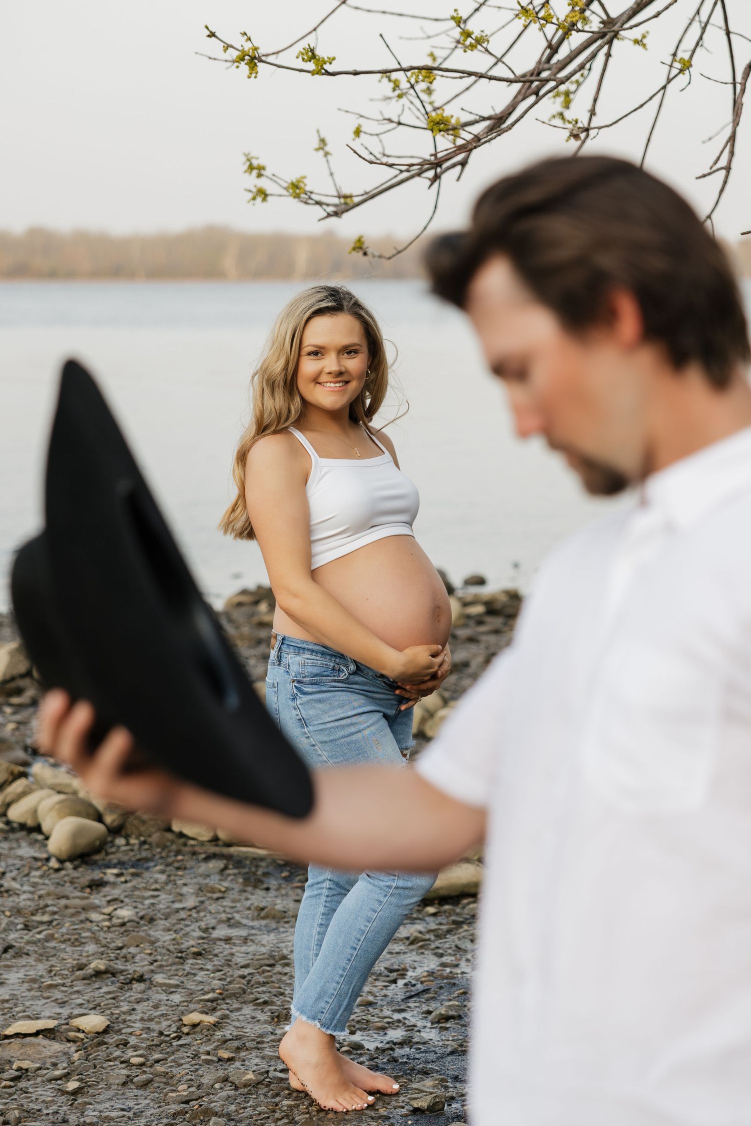Pregnant woman in white crop top and jeans smiling at man holding black cowboy hat by rocky riverside.