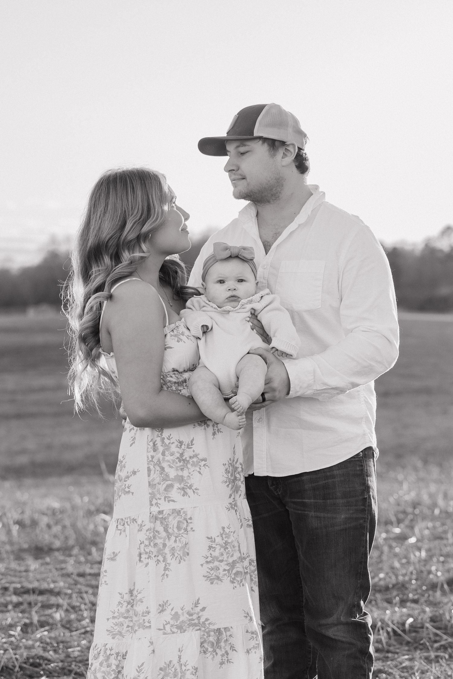 Black and white portrait of young parents holding their baby in an open field at sunset.