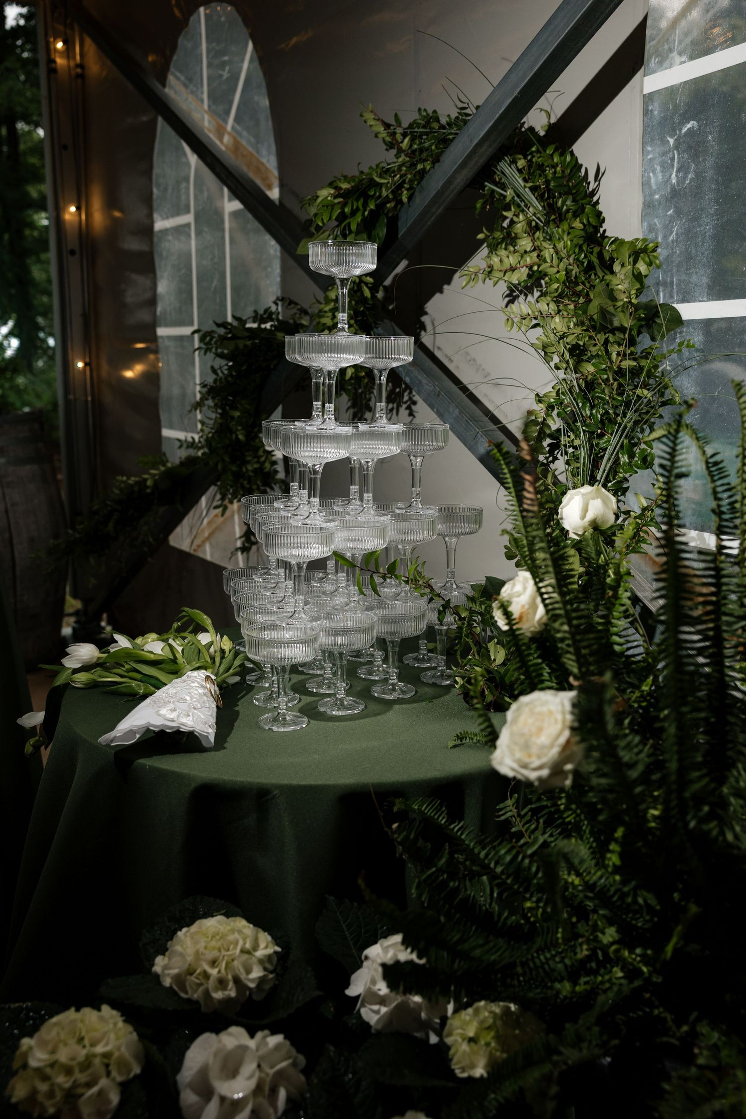 Champagne tower on green tablecloth surrounded by white roses, ferns, and greenery floral arch.