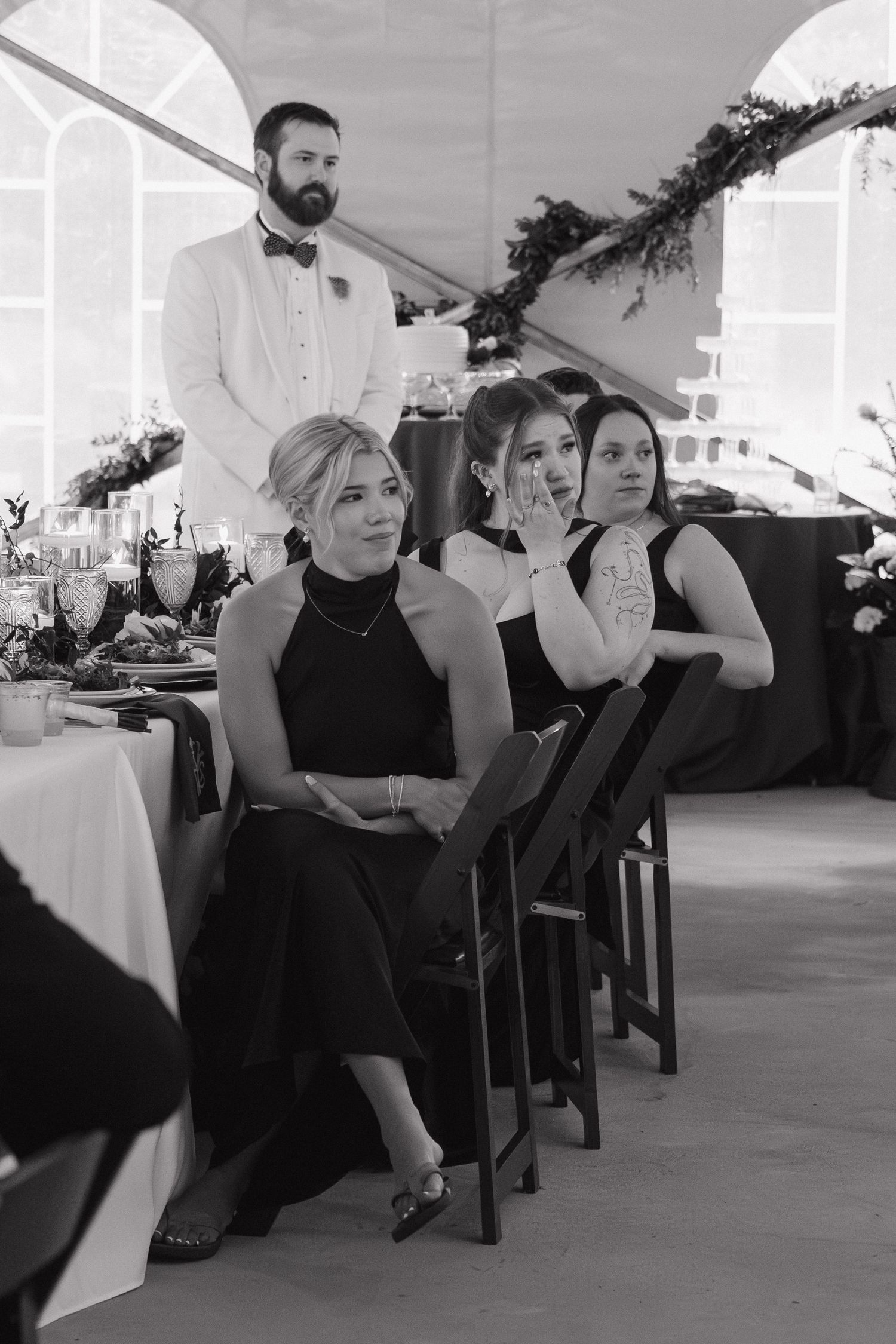 Black and white photo of bridesmaids in black dresses seated at wedding reception with groom standing behind.