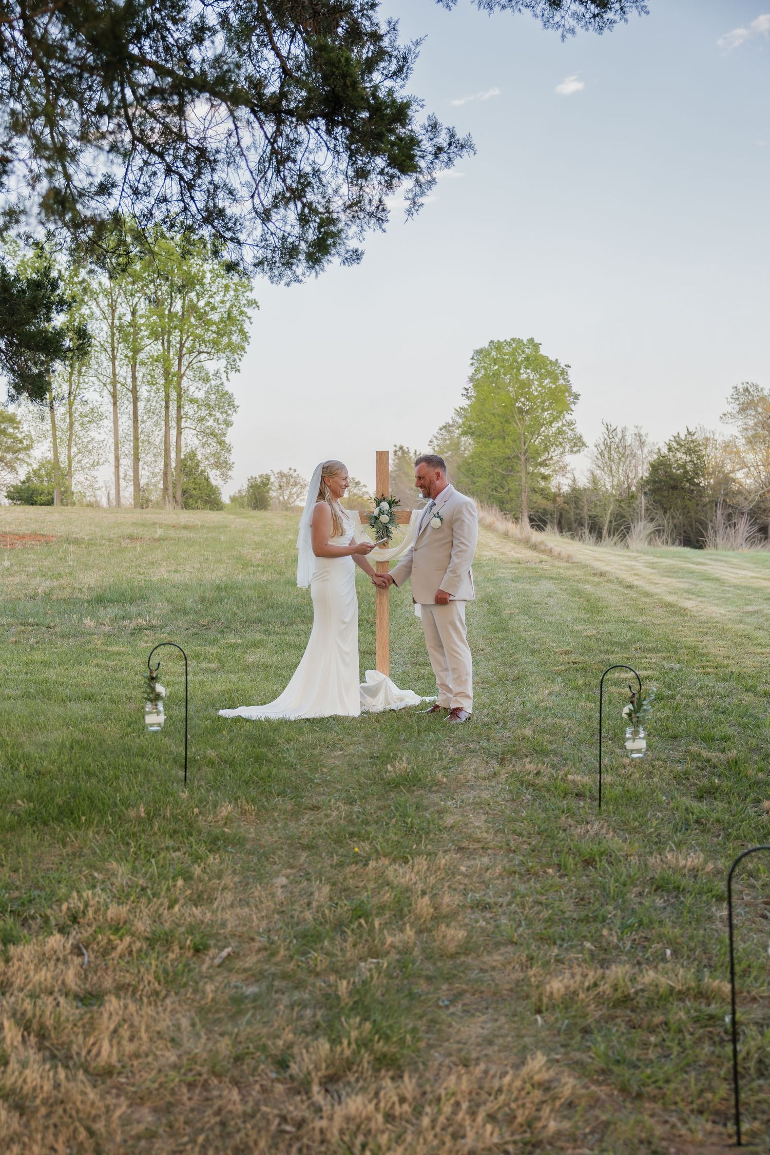 Bride and groom holding hands at rustic wooden cross altar during outdoor wedding ceremony.