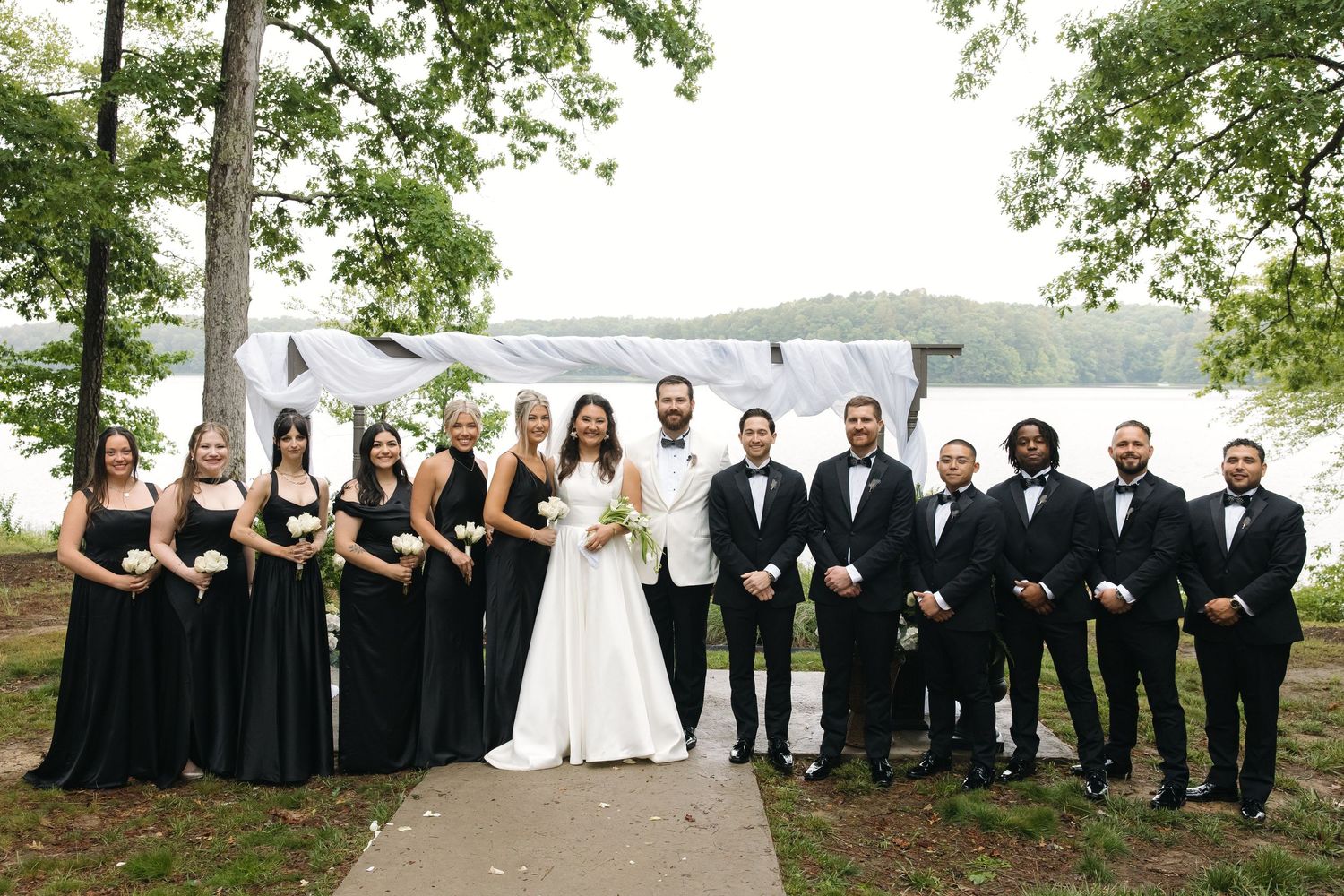 Wedding party poses lakeside with bridesmaids in black gowns and groomsmen in black tuxedos with bride and groom.