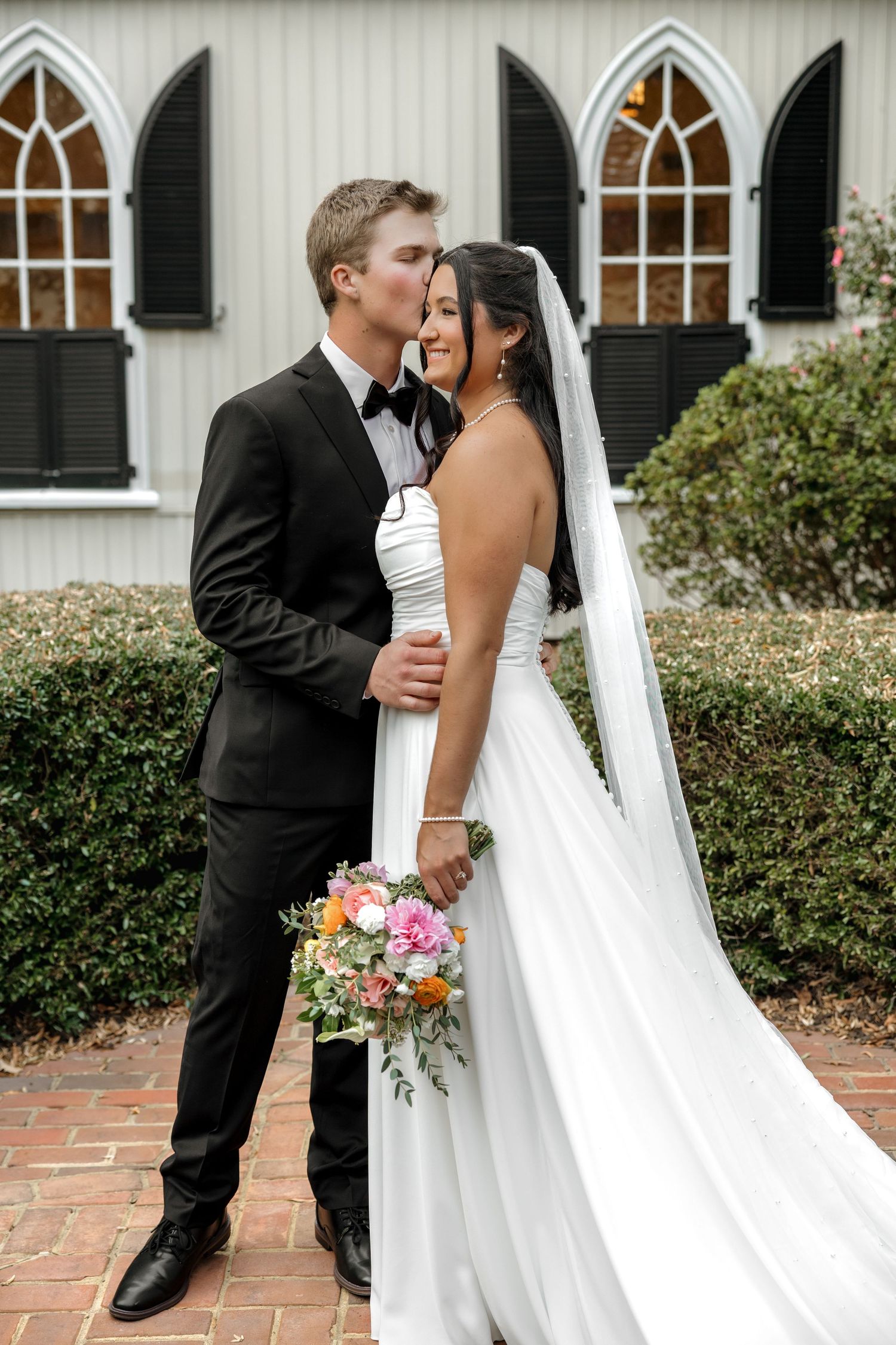 Groom kissing bride on cheek as she smiles, holding vibrant bouquet outside a white chapel.