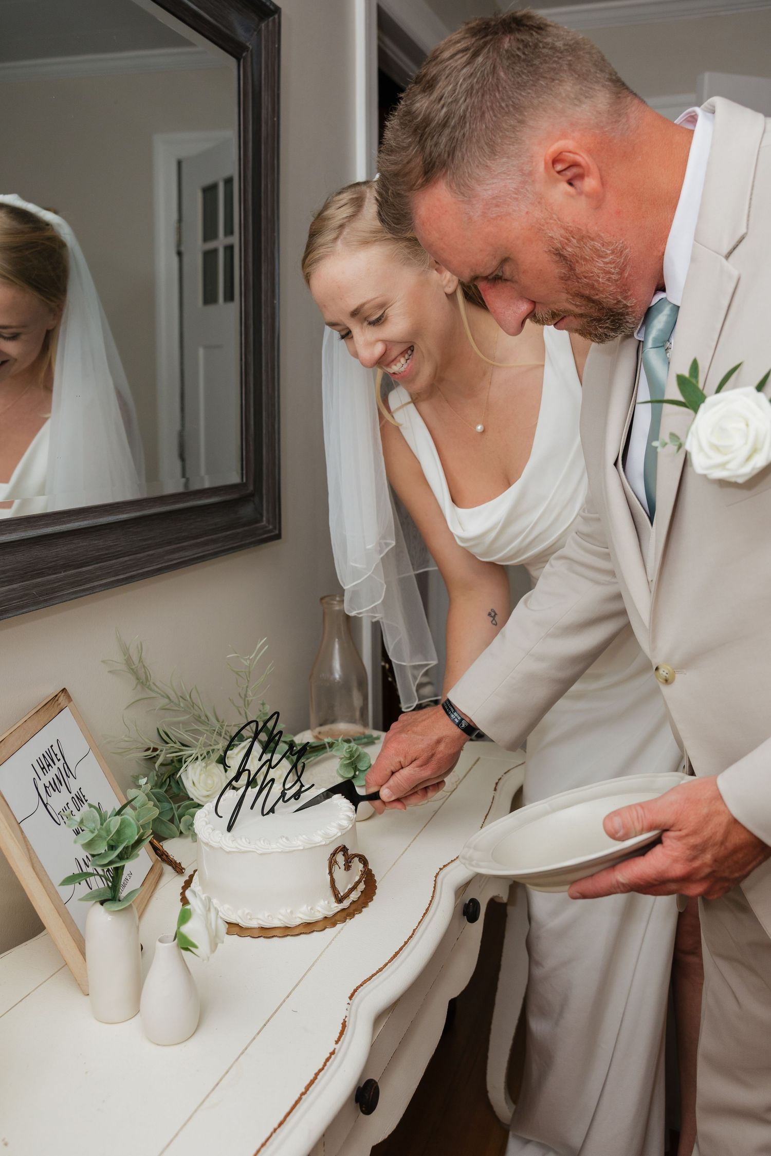 Smiling bride and groom cutting their white wedding cake decorated with Mr and Mrs topper and greenery.