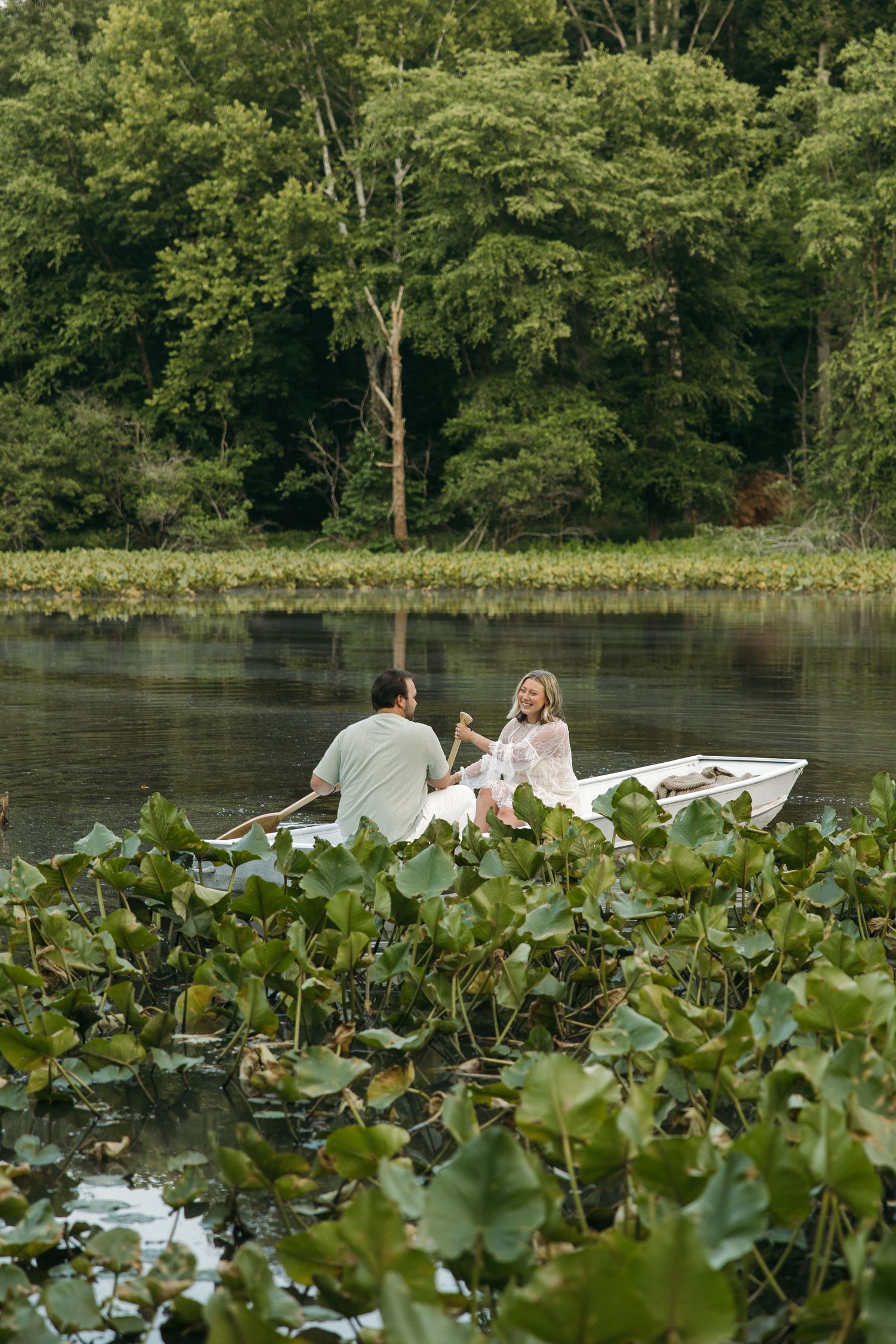 A smiling couple enjoys a romantic boat ride on a serene lake surrounded by green lily pads and forest.