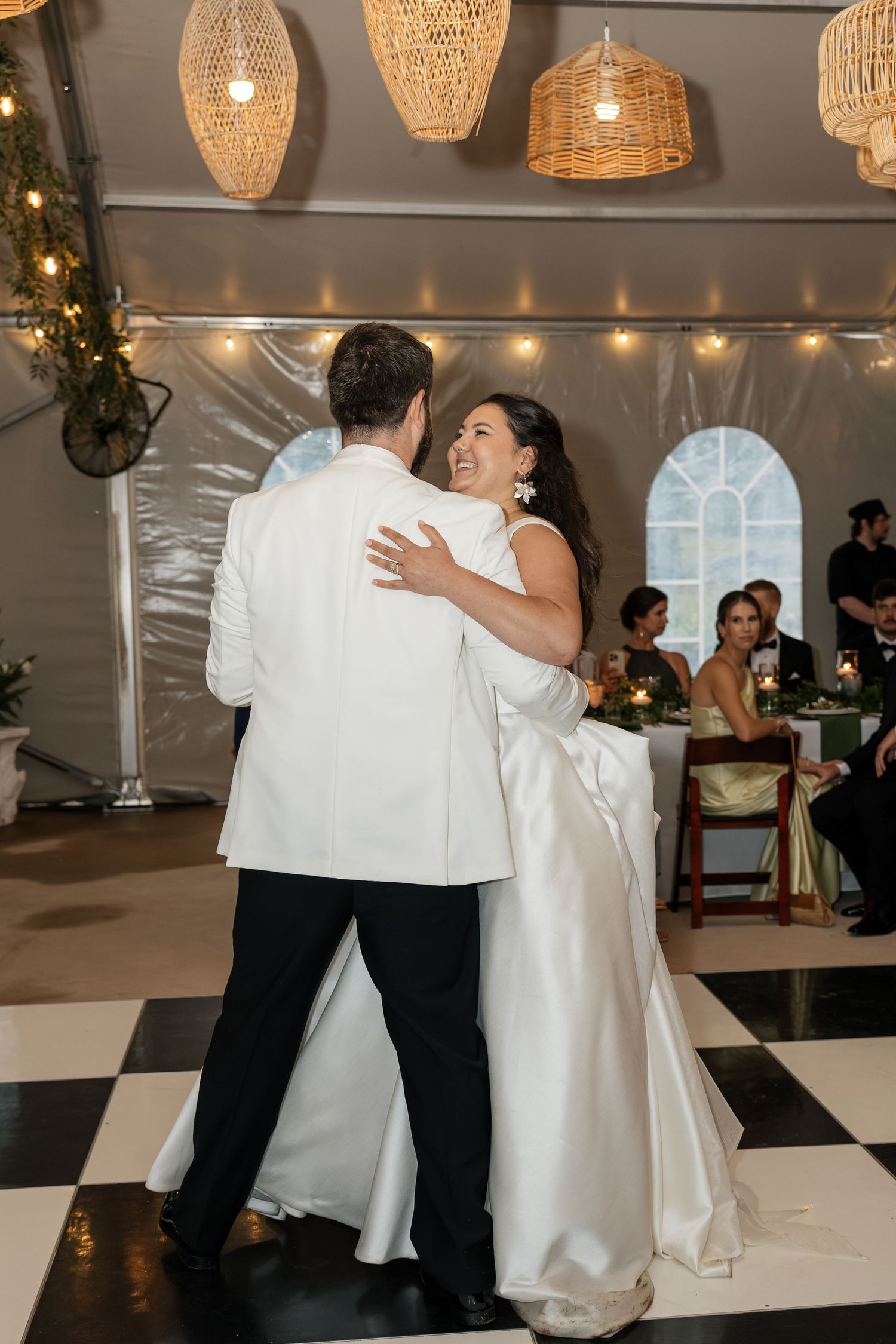 Bride and groom share first dance under wicker chandeliers on black and white checkered dance floor.