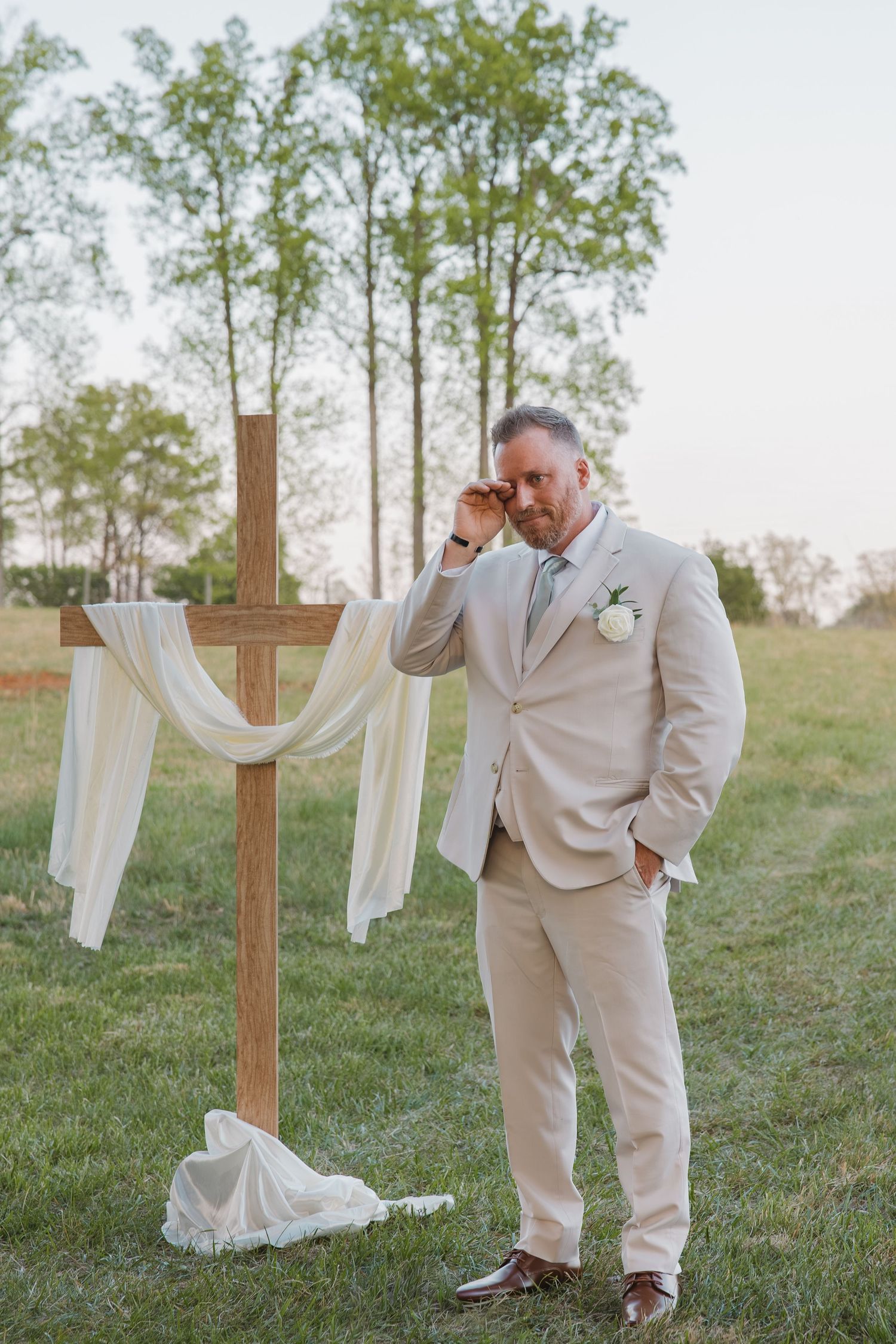 Emotional groom in beige suit wiping tear beside rustic wooden cross altar at outdoor wedding.