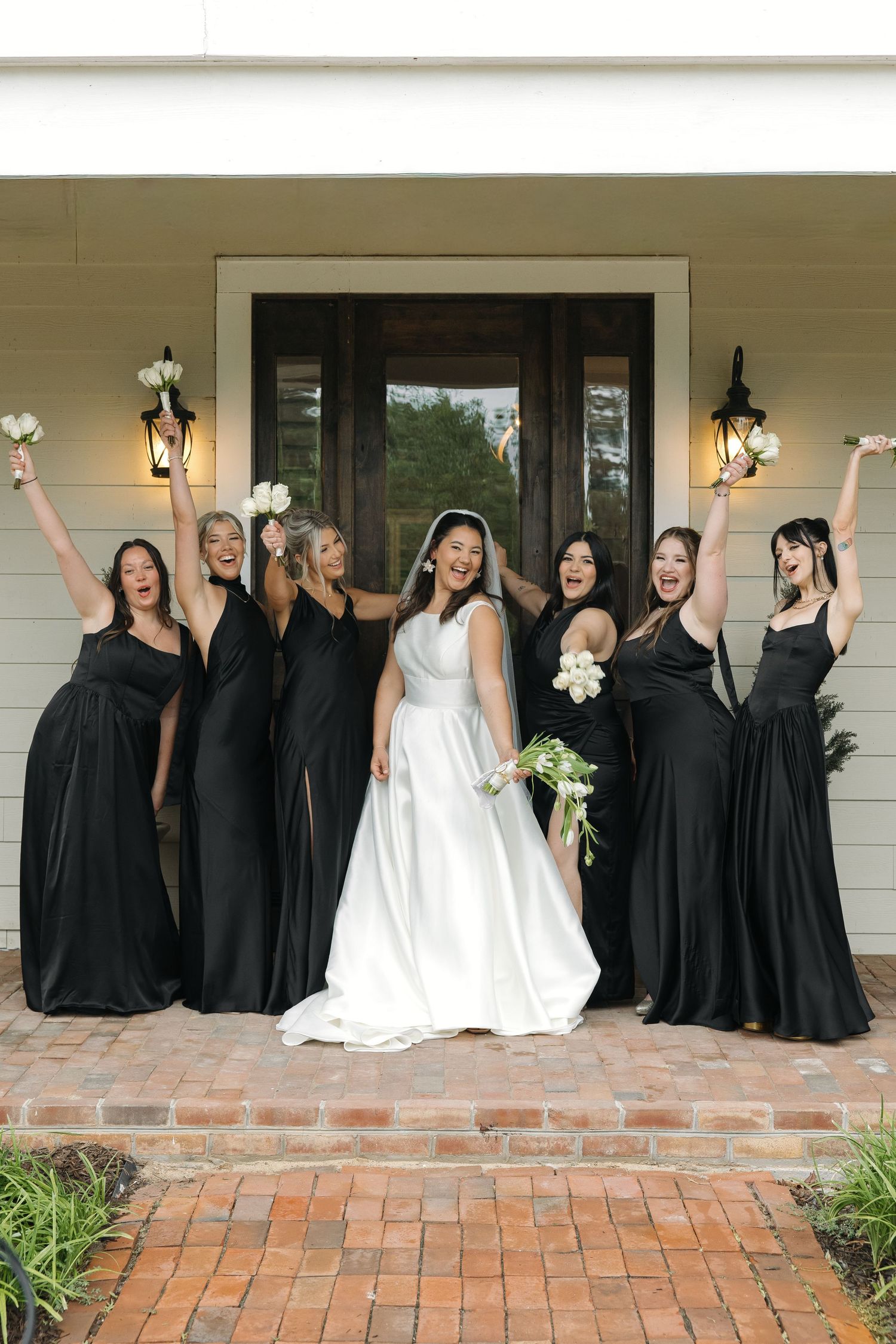 Joyful bride and bridesmaids in black gowns celebrate with raised arms outside a white home entrance.