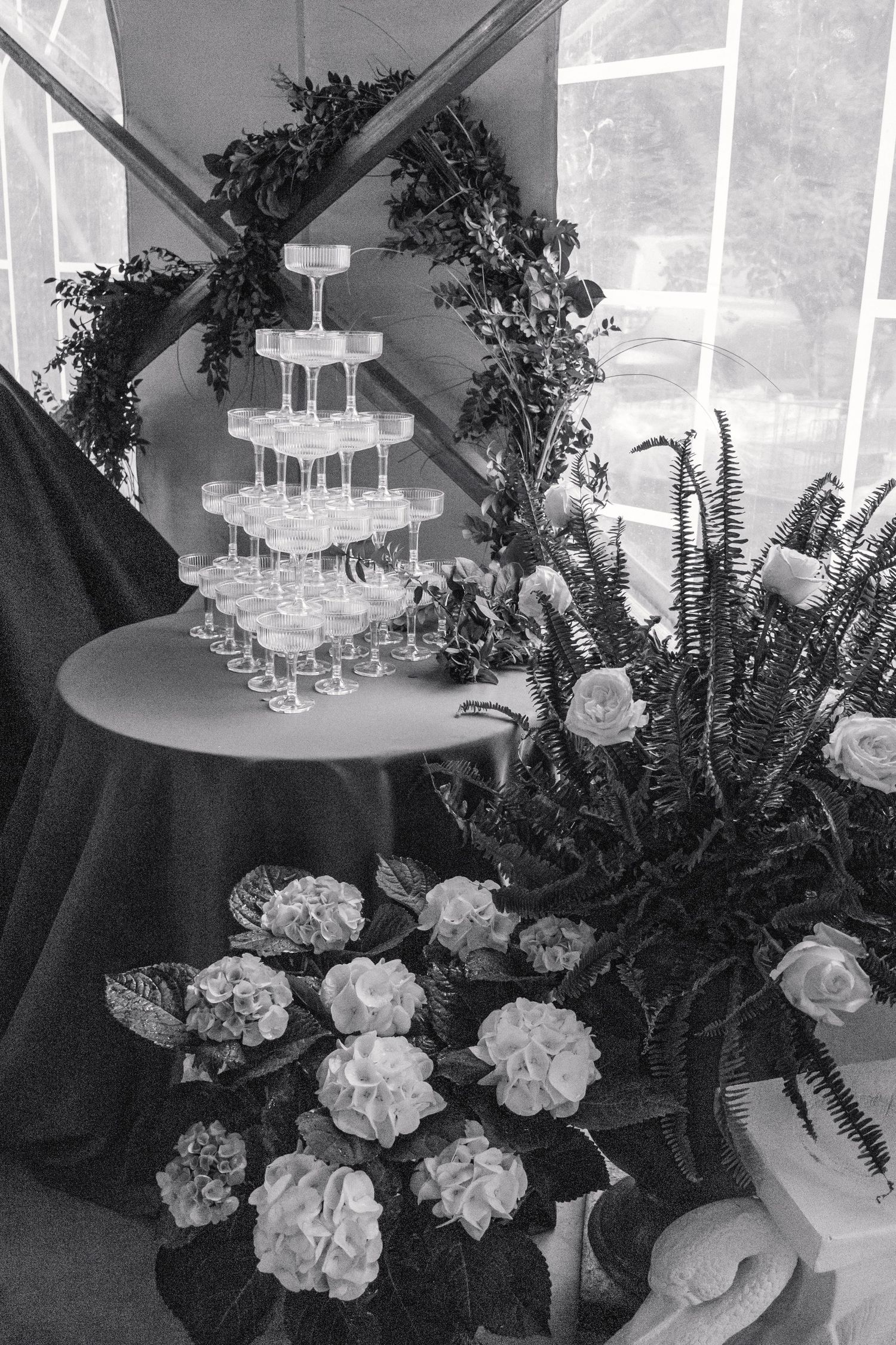 Black and white photo of champagne tower with floral arch and white flower arrangements at wedding.