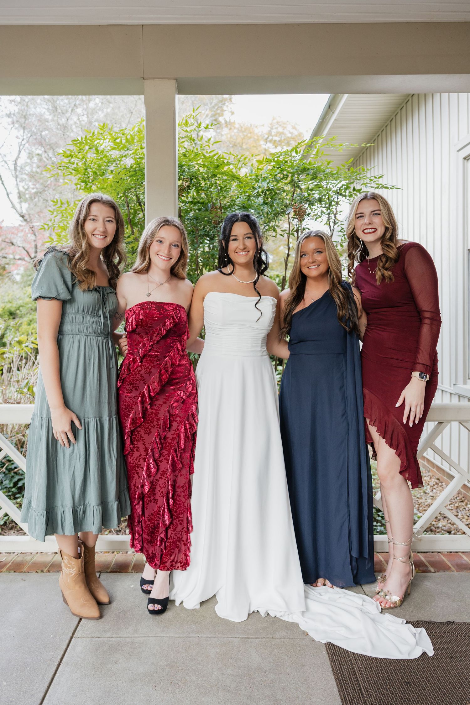 Bride in white gown smiling with four bridesmaids in colorful dresses on a covered porch.