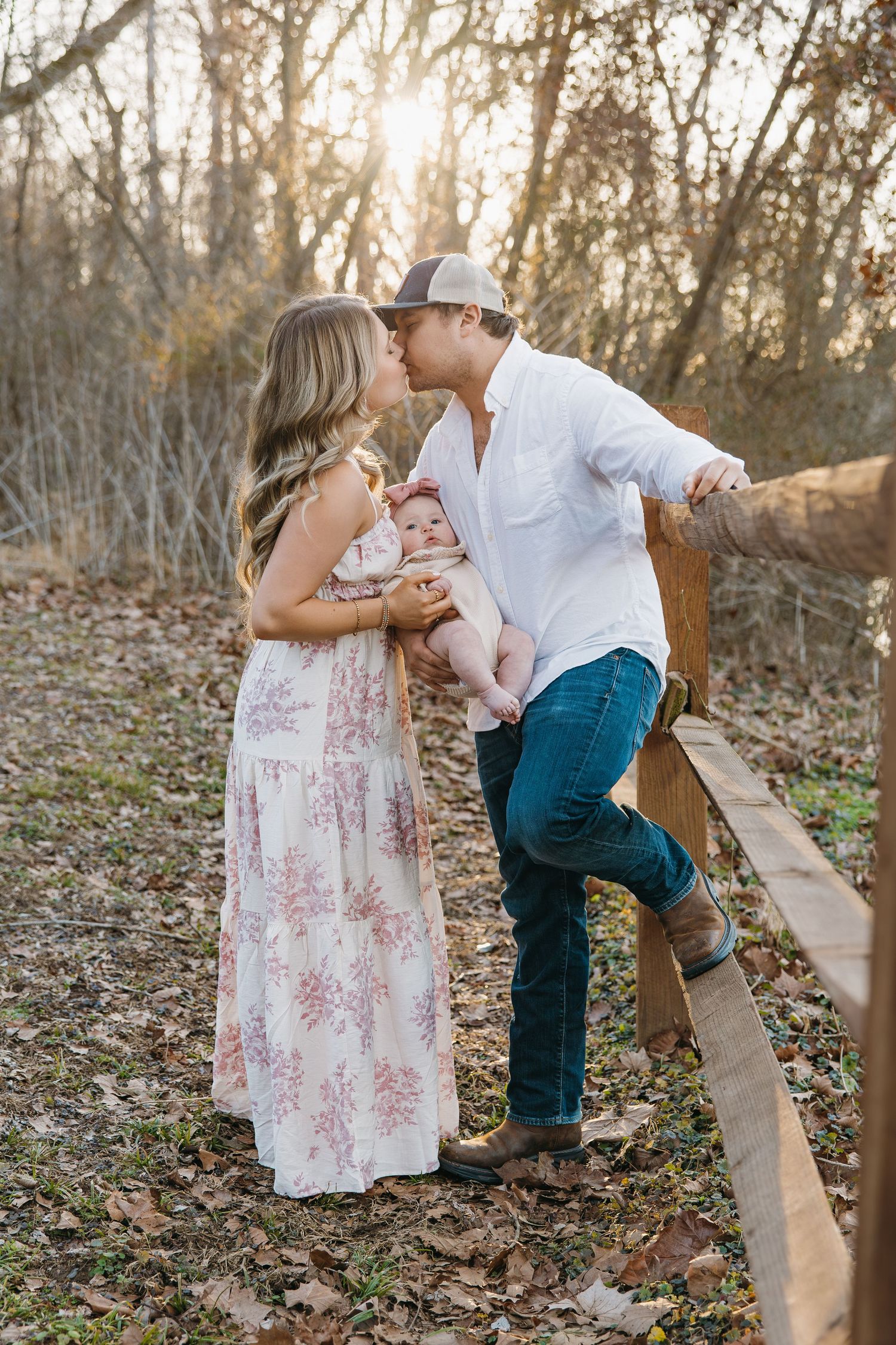 Young couple kissing outdoors at golden hour while holding their baby near a rustic wooden fence.