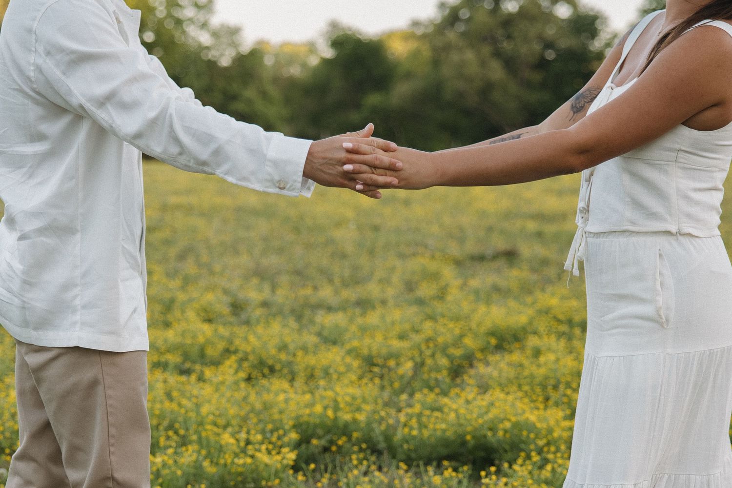 A couple holds hands while standing in a vibrant yellow wildflower field wearing white outfits.