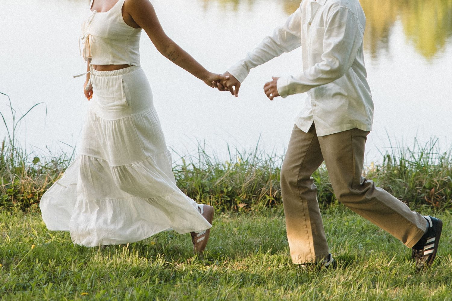Couple in white outfits running hand in hand along a grassy lakeside in warm golden sunlight.