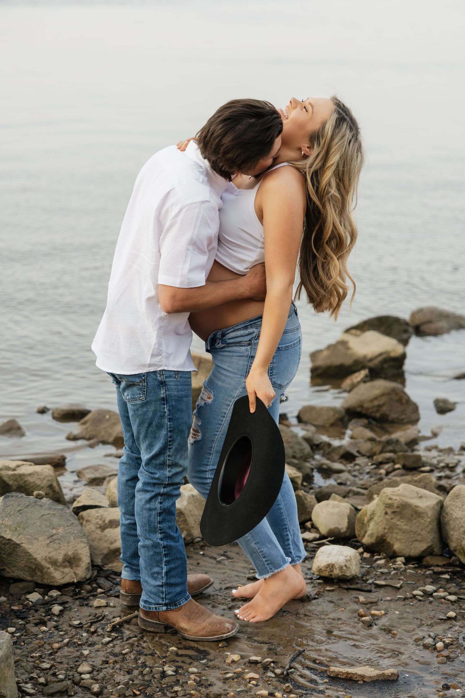 Man kissing laughing pregnant woman on rocky shoreline, woman holding black cowboy hat near the water.