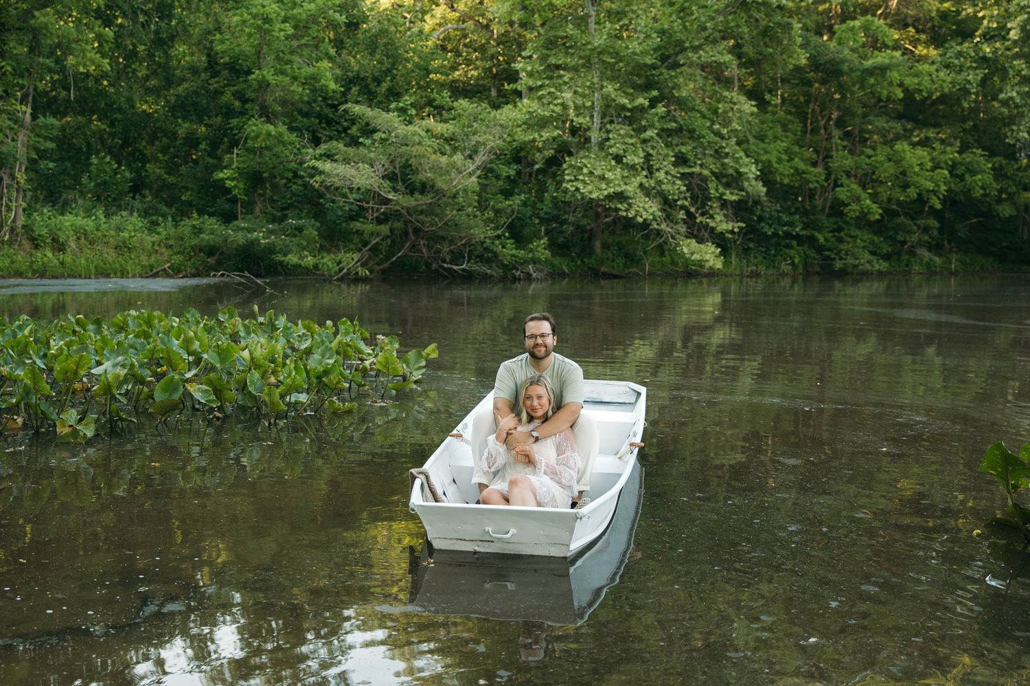 Happy couple embracing in a white rowboat floating on a peaceful tree-lined river.