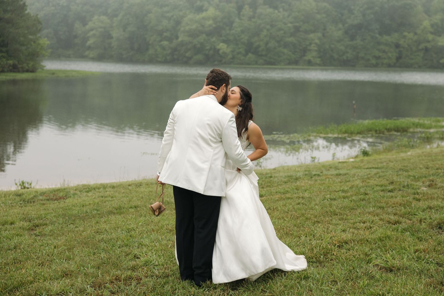 Bride and groom embrace in tender kiss near misty tree-lined lake backdrop.