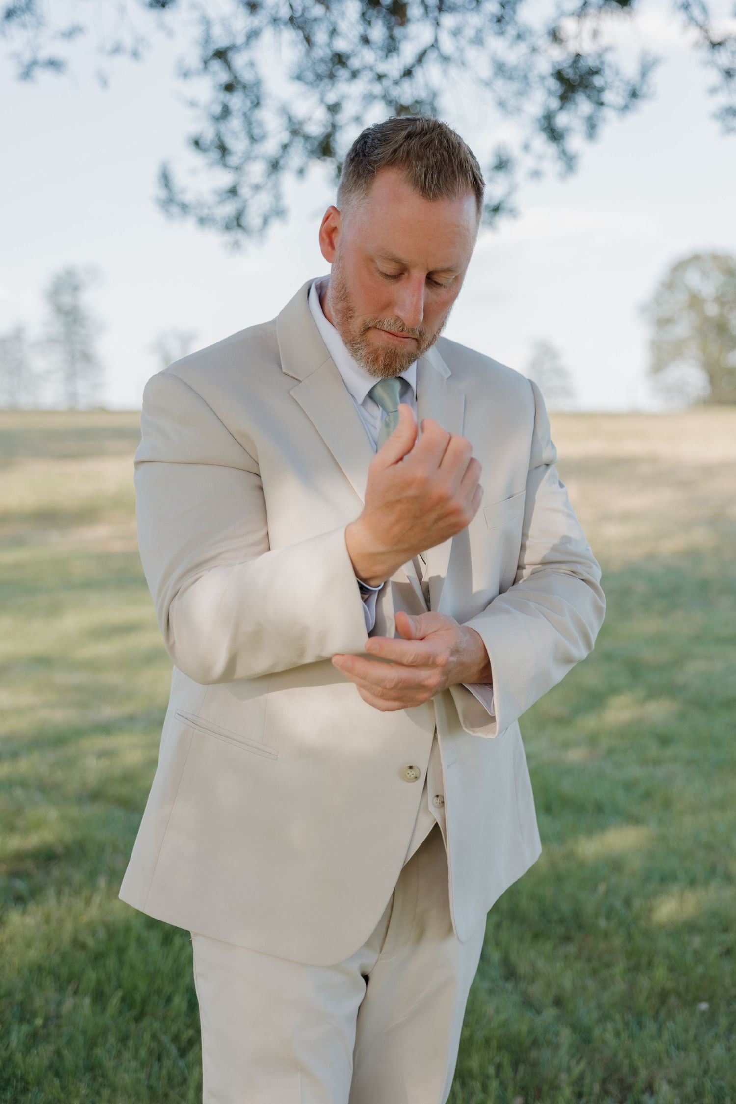 Groom in tan suit adjusting cufflinks outdoors in sunlit green field.