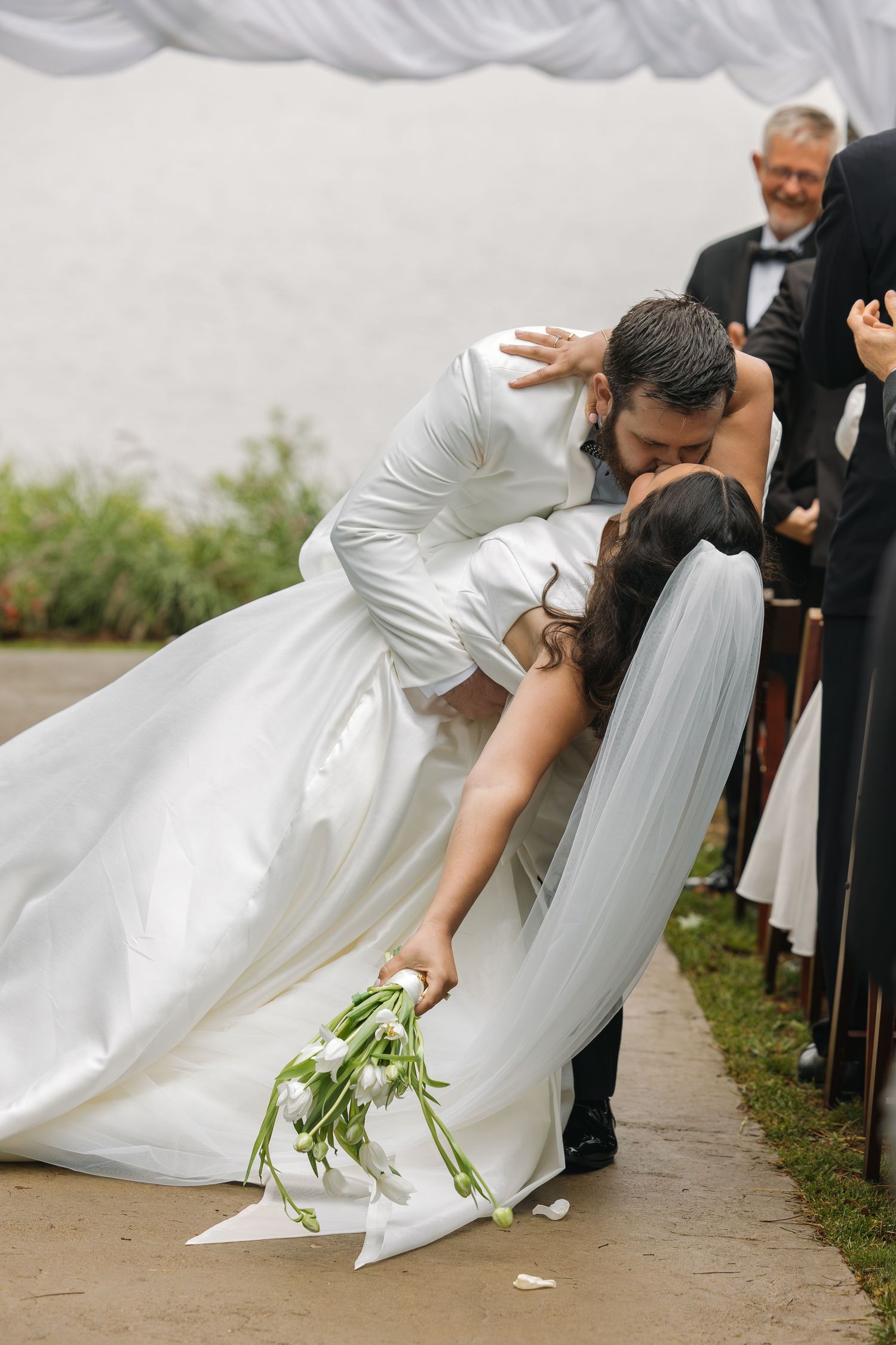Groom dips bride for a romantic kiss in the aisle after wedding ceremony, bride holding white tulip bouquet.