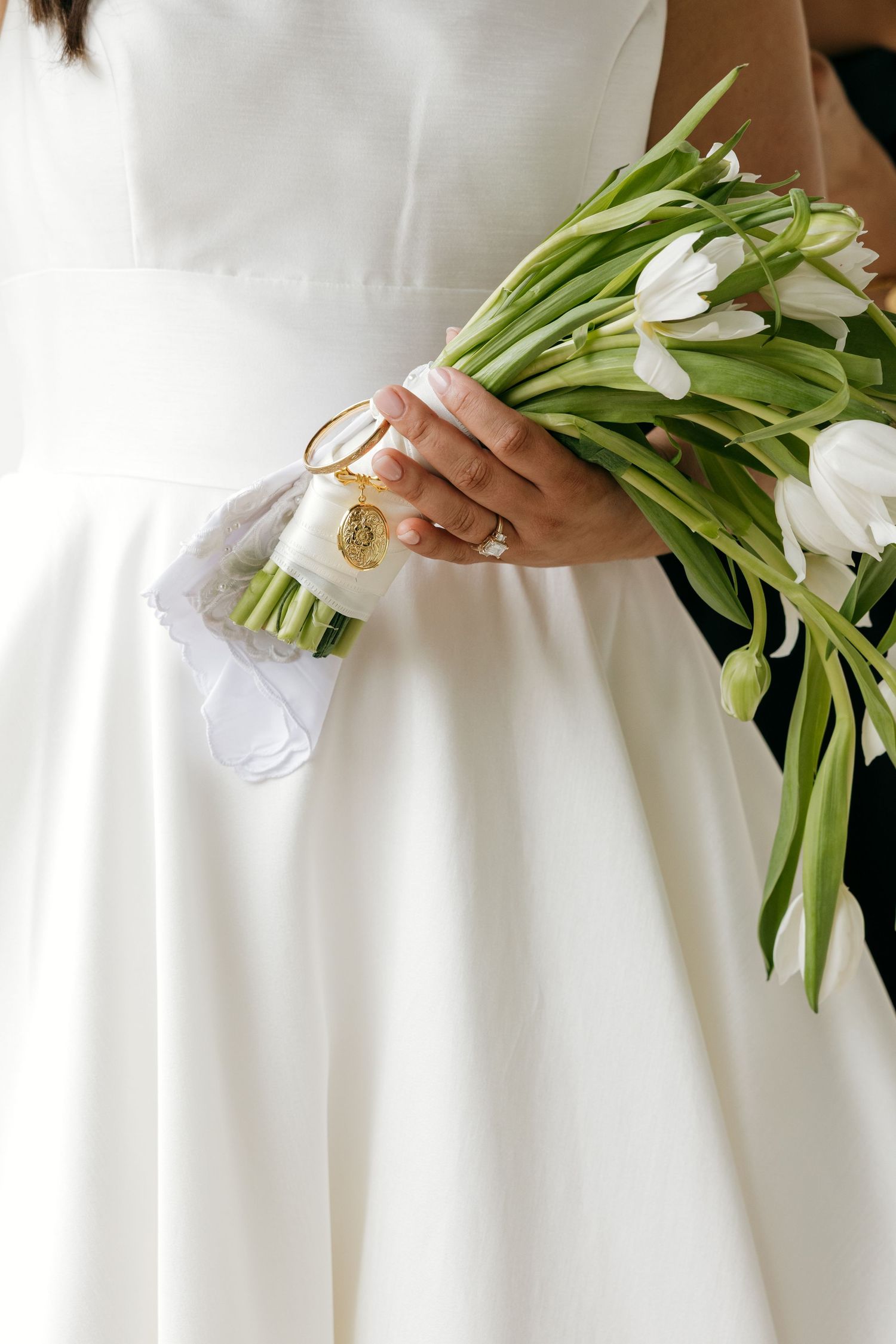 Bride in white satin gown holding a bouquet of white tulips with gold charm ribbon wrap detail.
