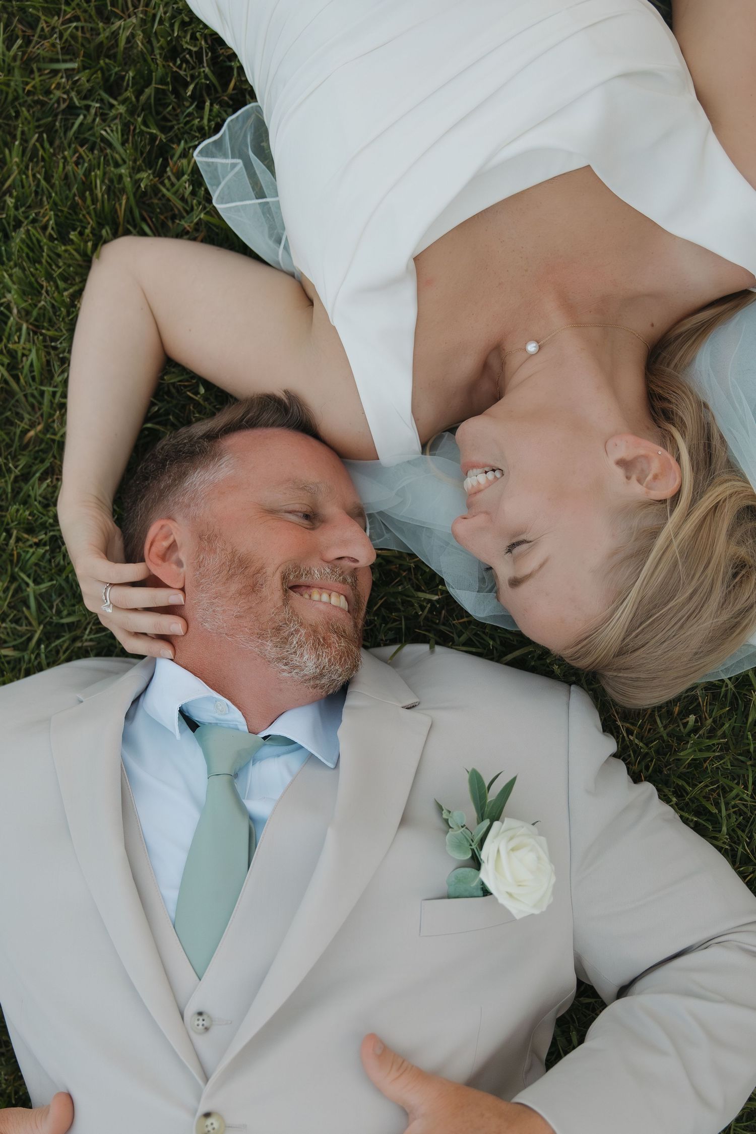 Bride and groom lay on green grass laughing together, groom wearing beige suit with white boutonniere.