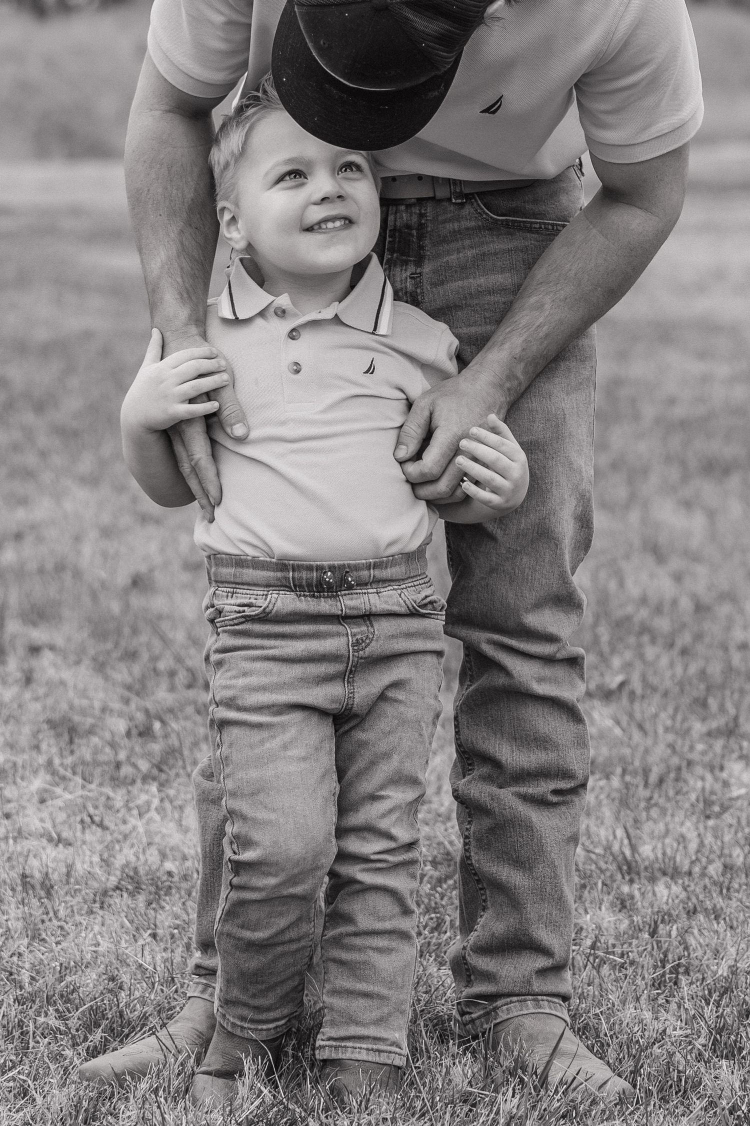 Black and white photo of a smiling young boy standing barefoot in grass while his father holds him from behind.