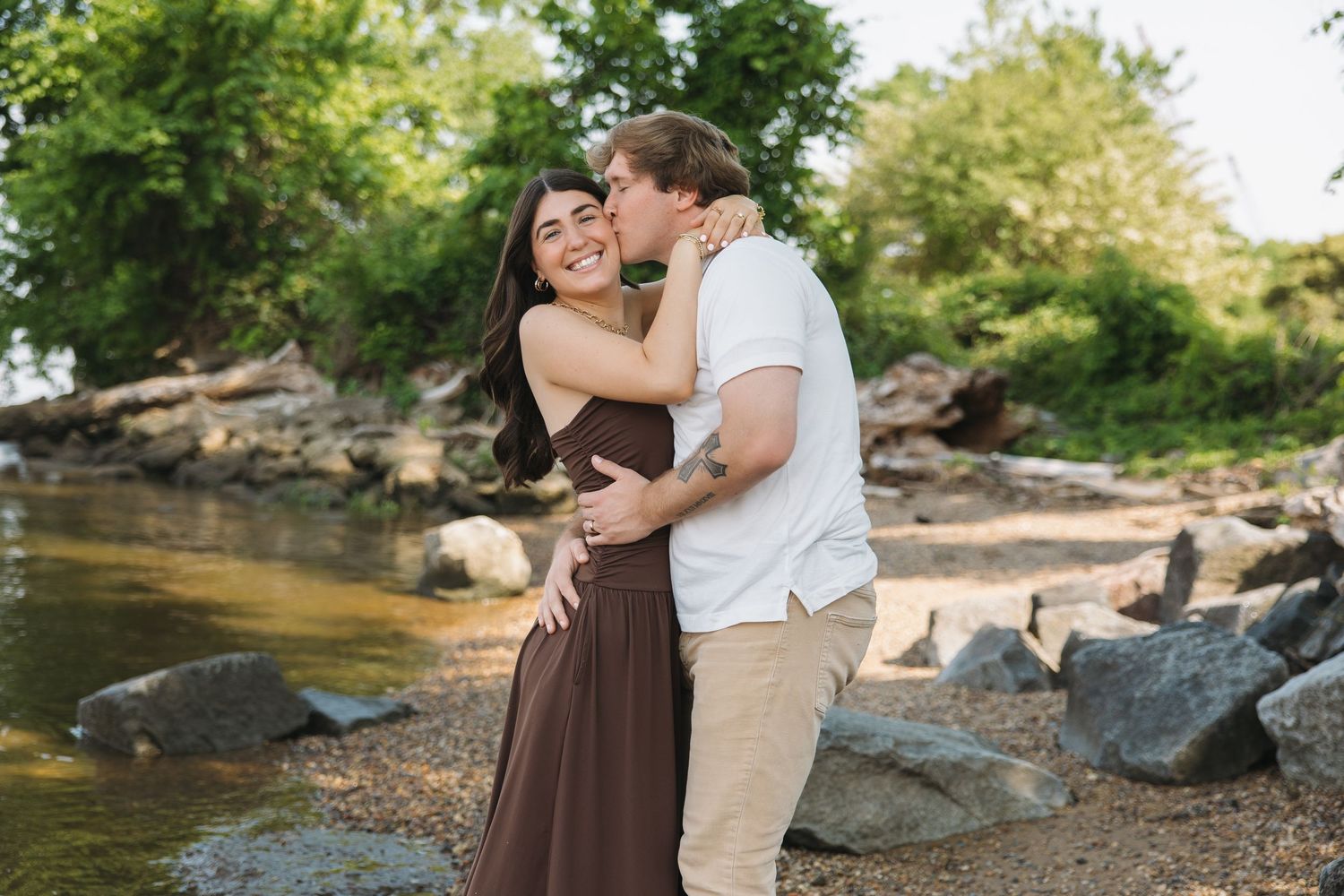 A smiling couple embraces near a rocky riverside, the man kissing her cheek in warm natural light.