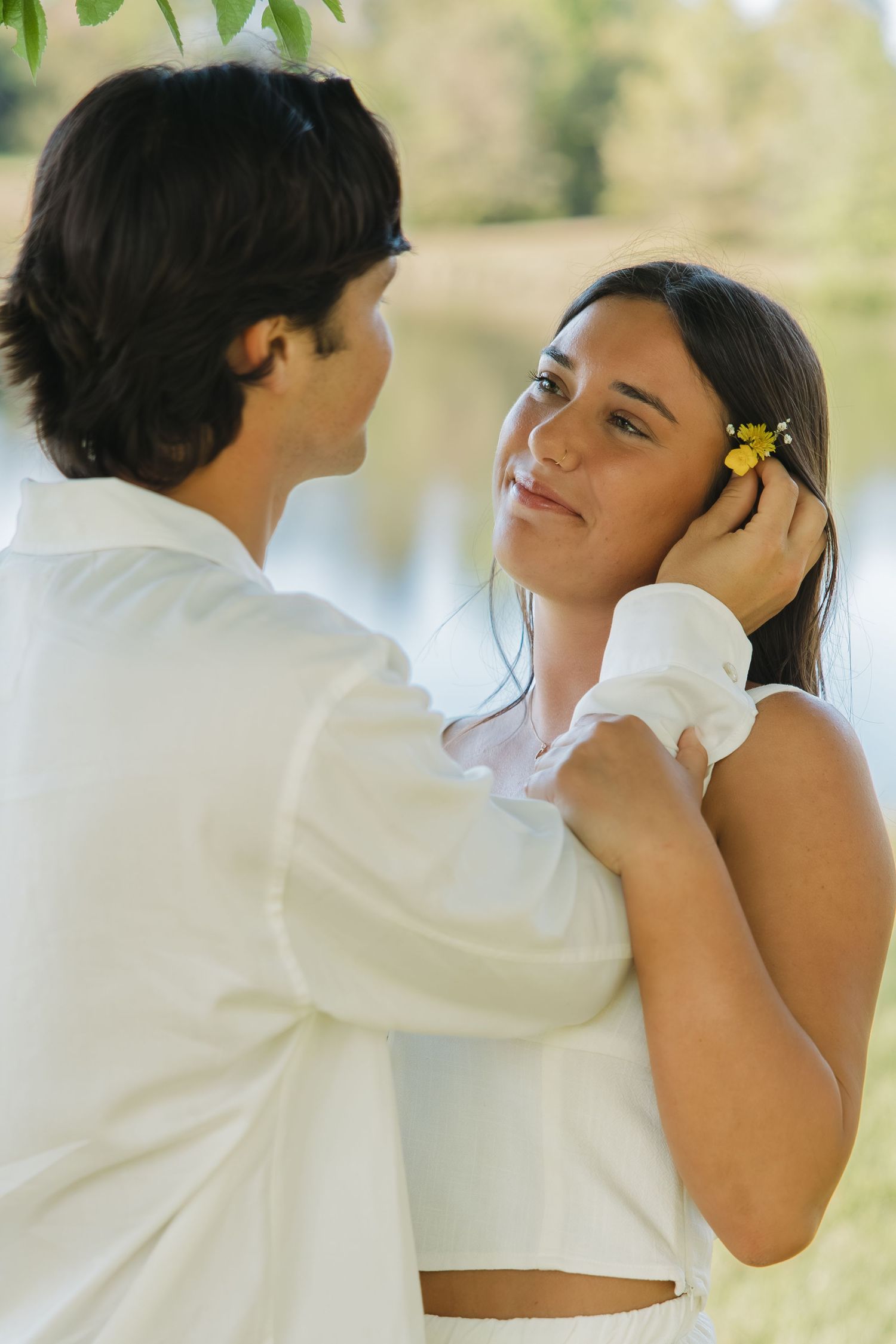 Romantic couple in white outfits sharing an intimate moment as man tucks flower into woman's hair by a lake.