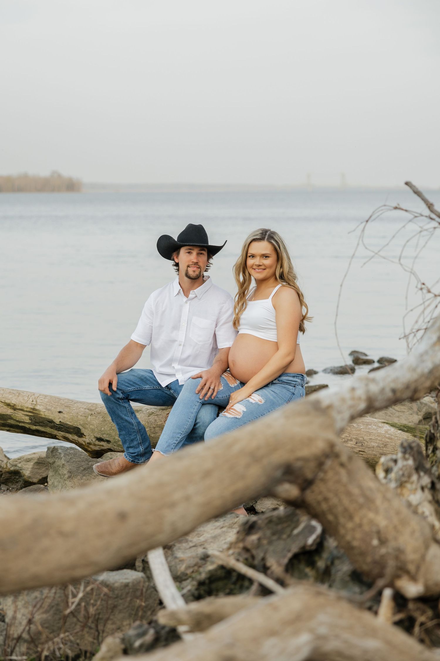Expecting couple sitting on driftwood by the water, man in cowboy hat embracing pregnant woman in white crop top.