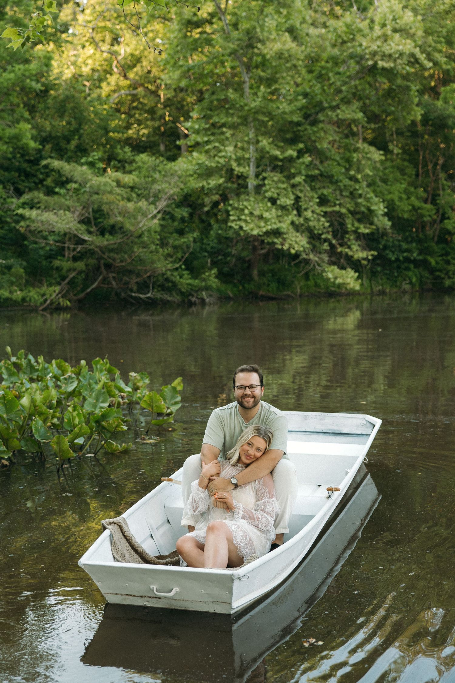 Couple embracing in a white rowboat on a serene tree-lined river during golden hour.