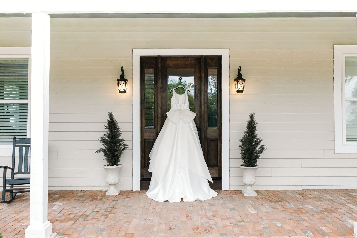 White wedding gown hanging on a charming farmhouse porch with brick flooring and lantern wall sconces.