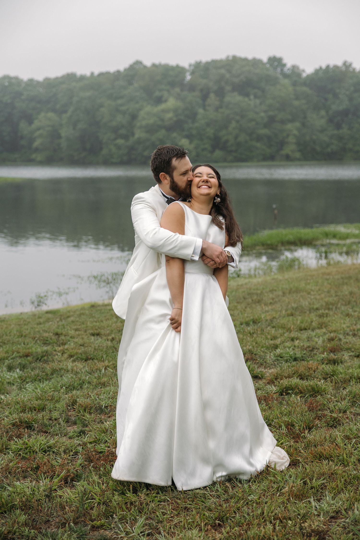 Groom tenderly kisses bride's cheek as they embrace near misty lake backdrop.