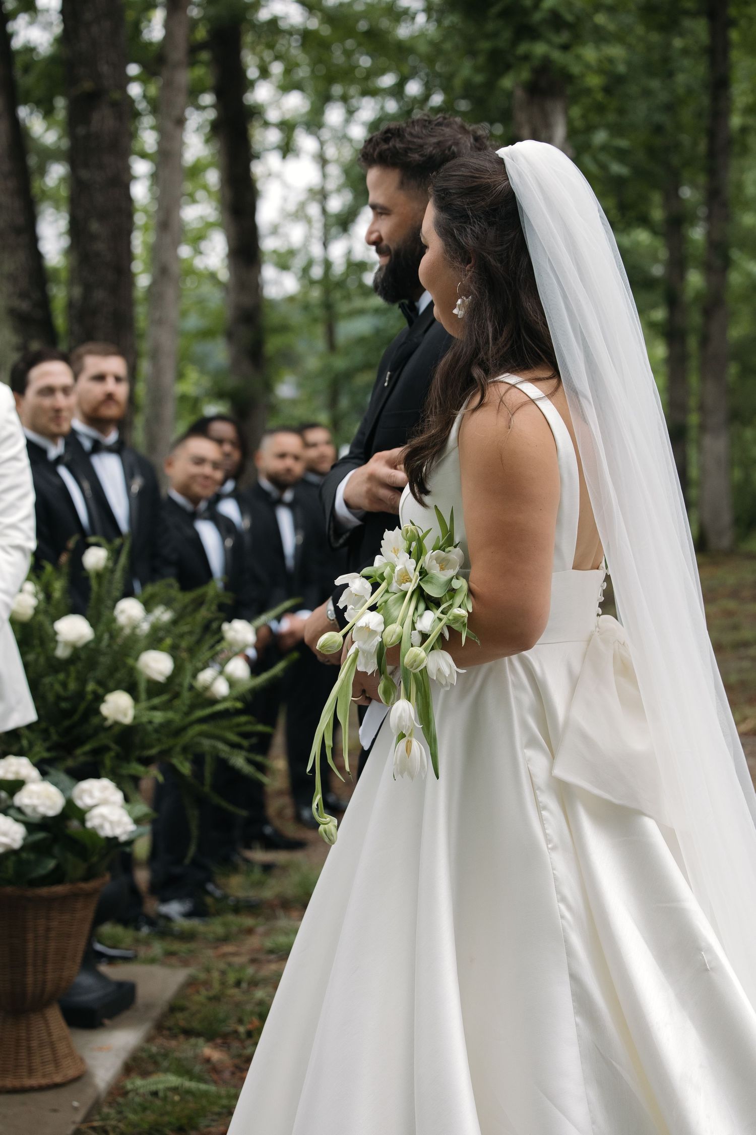 Bride in white gown with veil holds tulip bouquet beside groom in tuxedo at outdoor forest wedding ceremony.