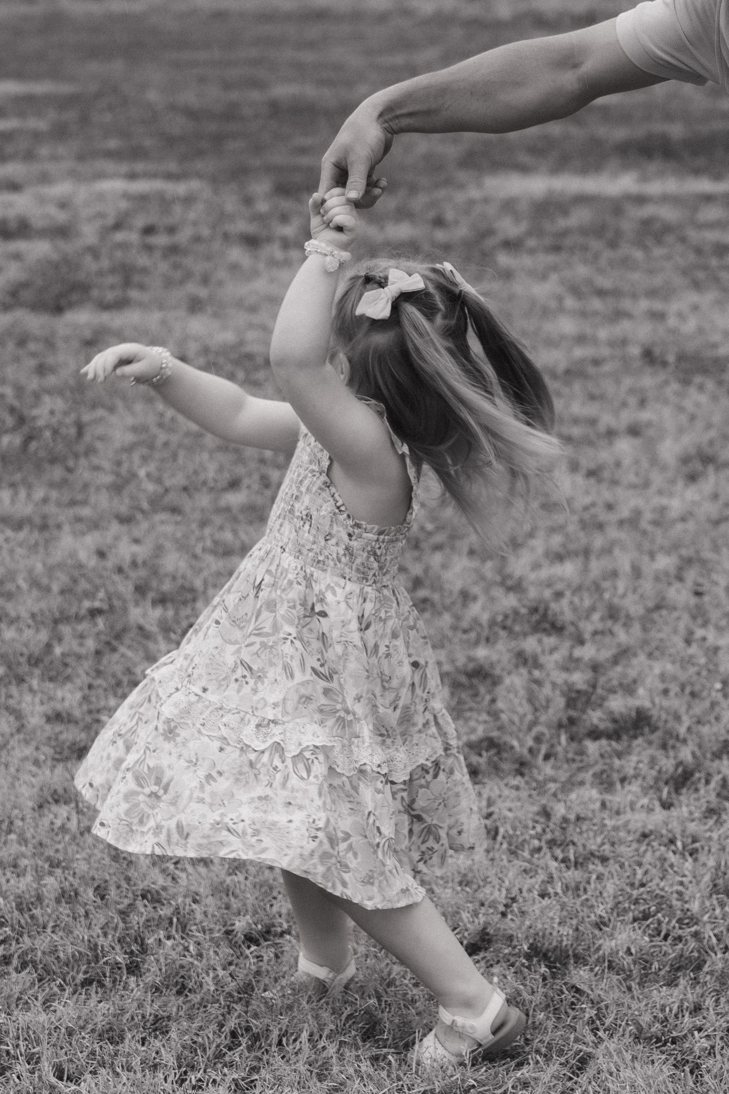 Black and white photo of a young girl in a floral dress twirling while holding an adults hand on a grass field.
