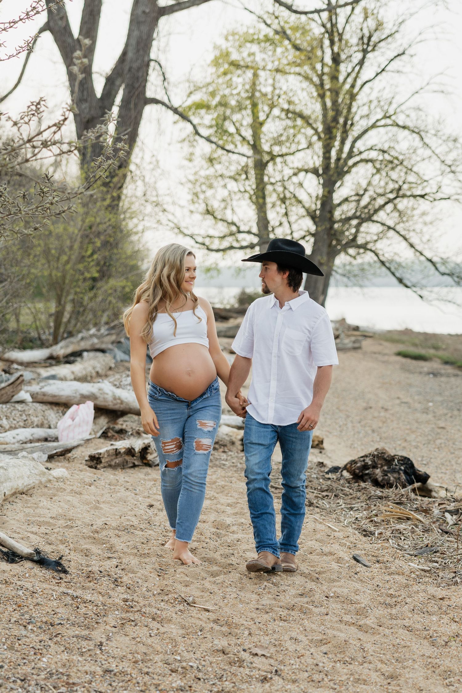 Pregnant couple walks hand in hand along sandy beach path, man in black cowboy hat and woman in ripped jeans.
