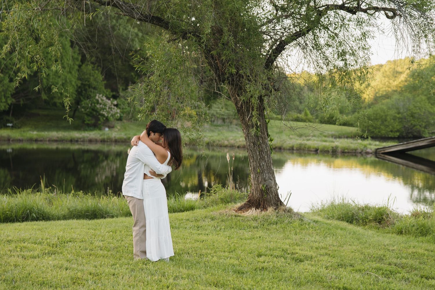 Romantic couple sharing a kiss beside a tranquil lake under a large willow tree outdoors.