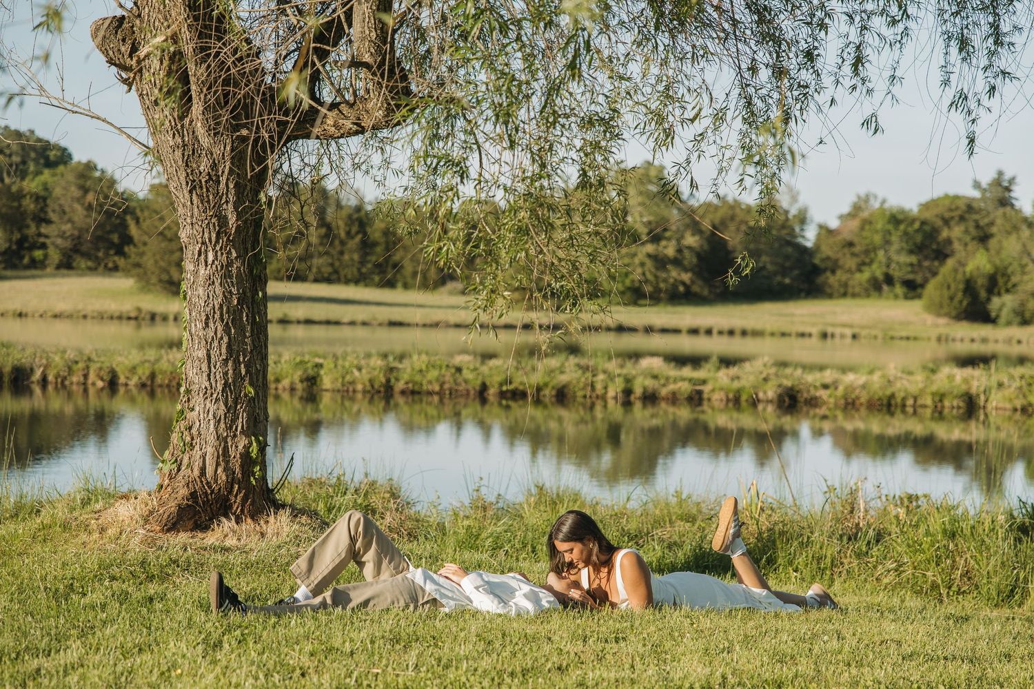 Couple relaxing side by side on grass under a large tree beside a serene lake on a bright sunny day.