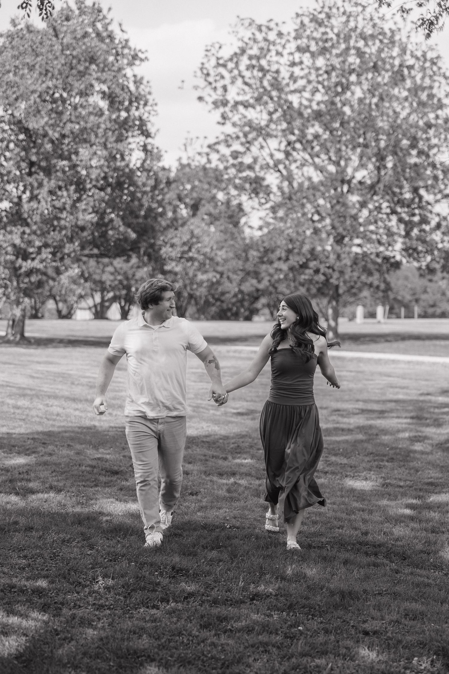 Black and white photo of a happy couple holding hands and walking through a grassy park together.