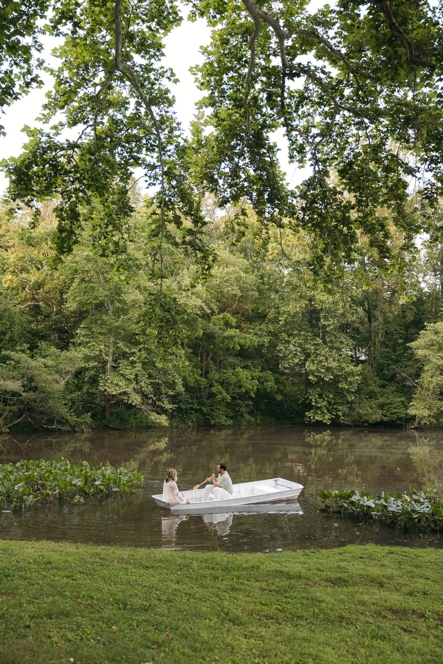 Two people sitting in a white rowboat on a calm river surrounded by lush green trees.