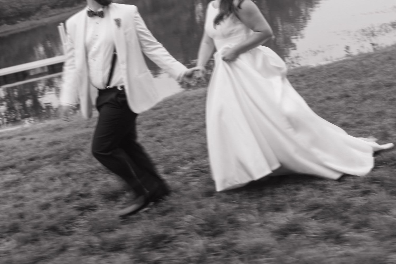 Black and white photo of bride and groom holding hands running by lakeside.