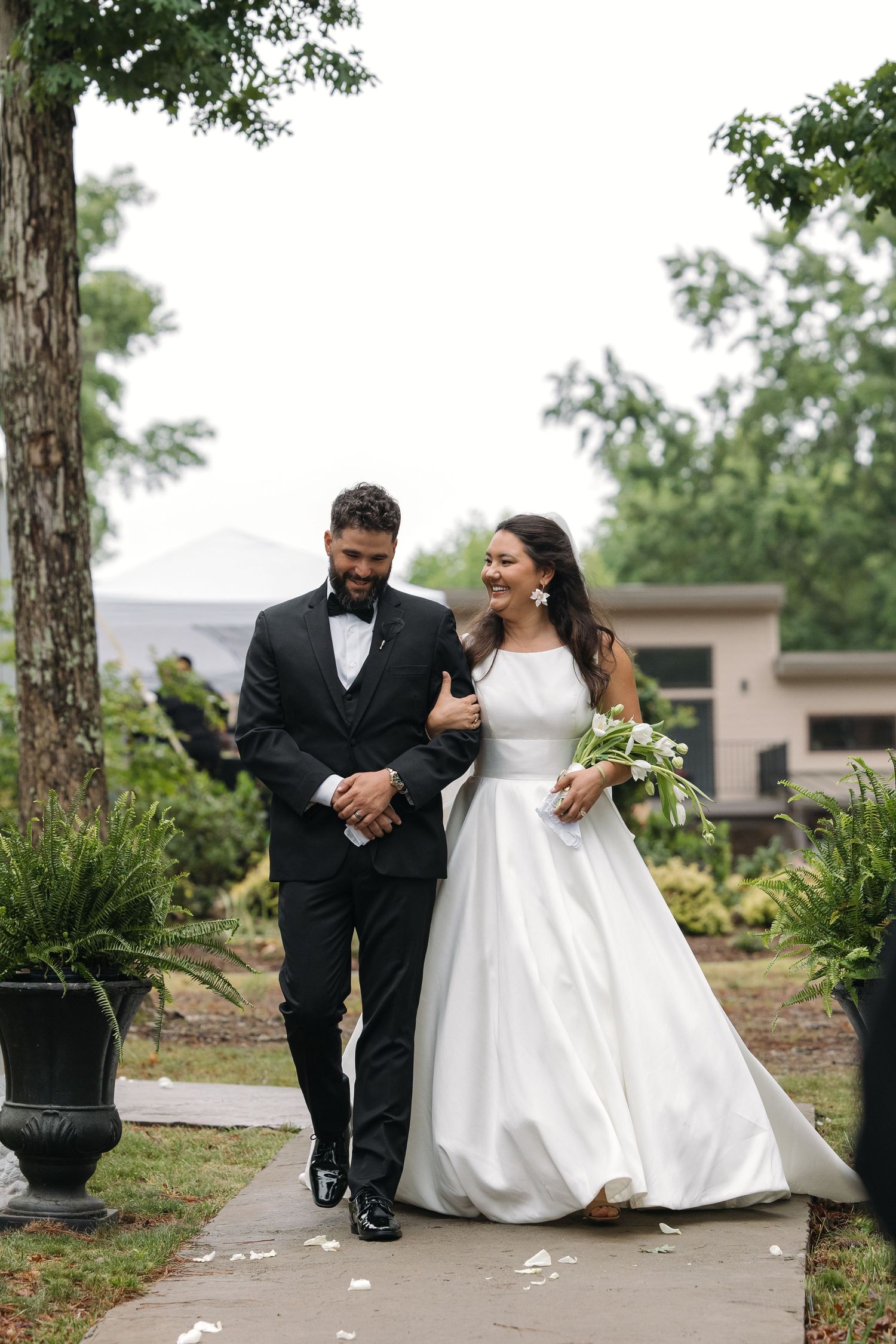 Bride in white gown and groom in black tuxedo walking joyfully down outdoor wedding aisle.