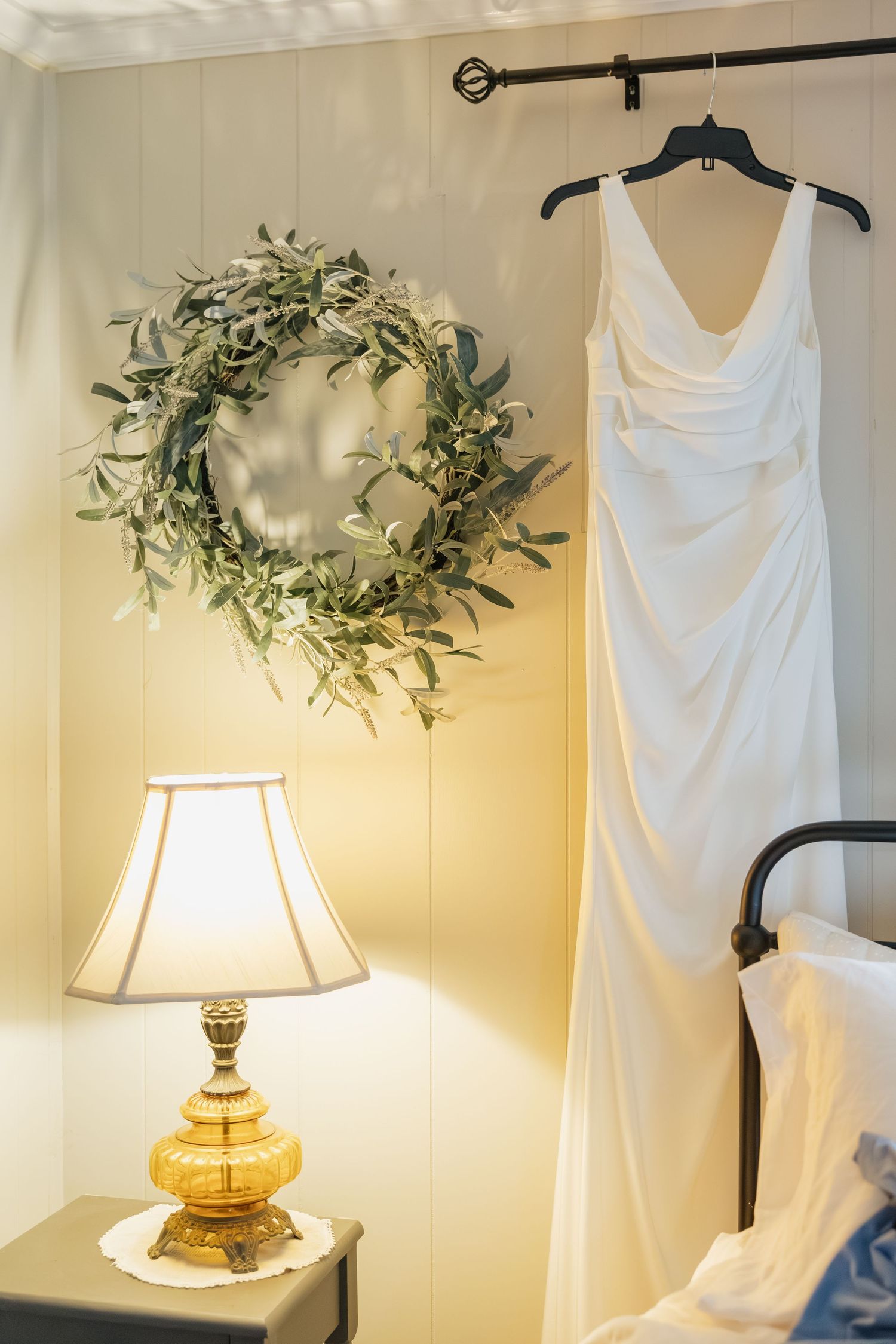 White wedding gown hanging near olive wreath and glowing lamp in cozy bridal suite.