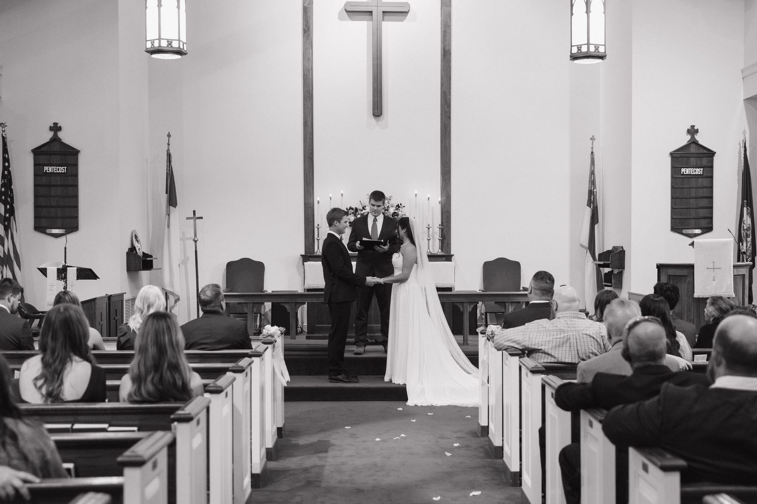 Black and white photo of bride and groom exchanging vows at church altar before seated guests.
