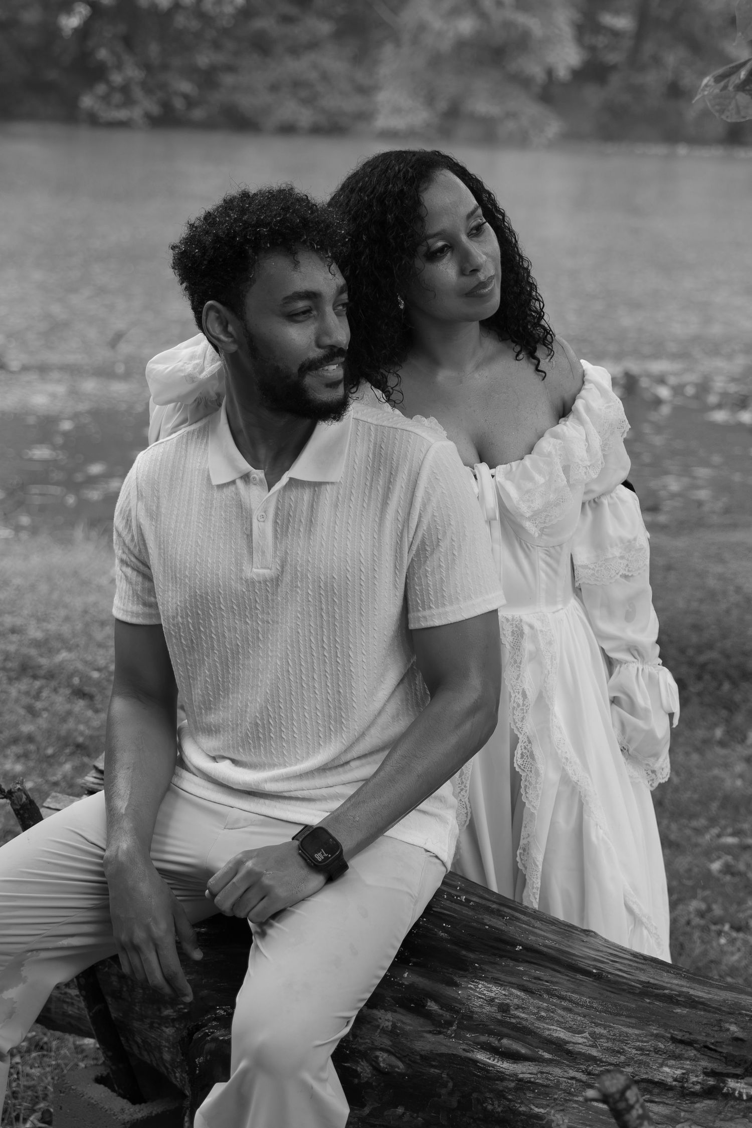 Black and white photo of a couple in white outfits posing near a log outdoors by a lake.